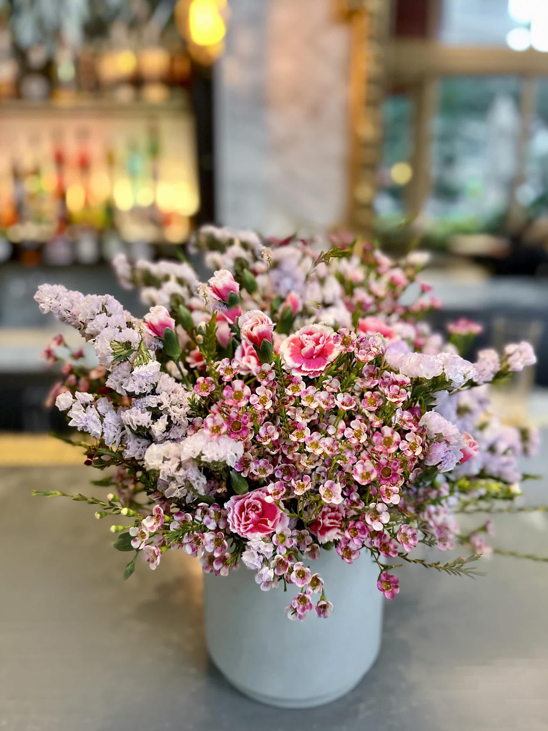 Pastel Roses Delight Vase - The &quot;Pastel Roses Delight Vase&quot; is a lovely arrangement featuring a blend of delicate pink and lavender flowers. This charming display brings a touch of whimsy and elegance to any space, making it a perfect choice for various occasions.  Arrangement Details: Main Flowers: Pink Spray Roses and Statice Container: Classic round white vase Approximate Dimensions: 18&quot;W x 16&quot;H