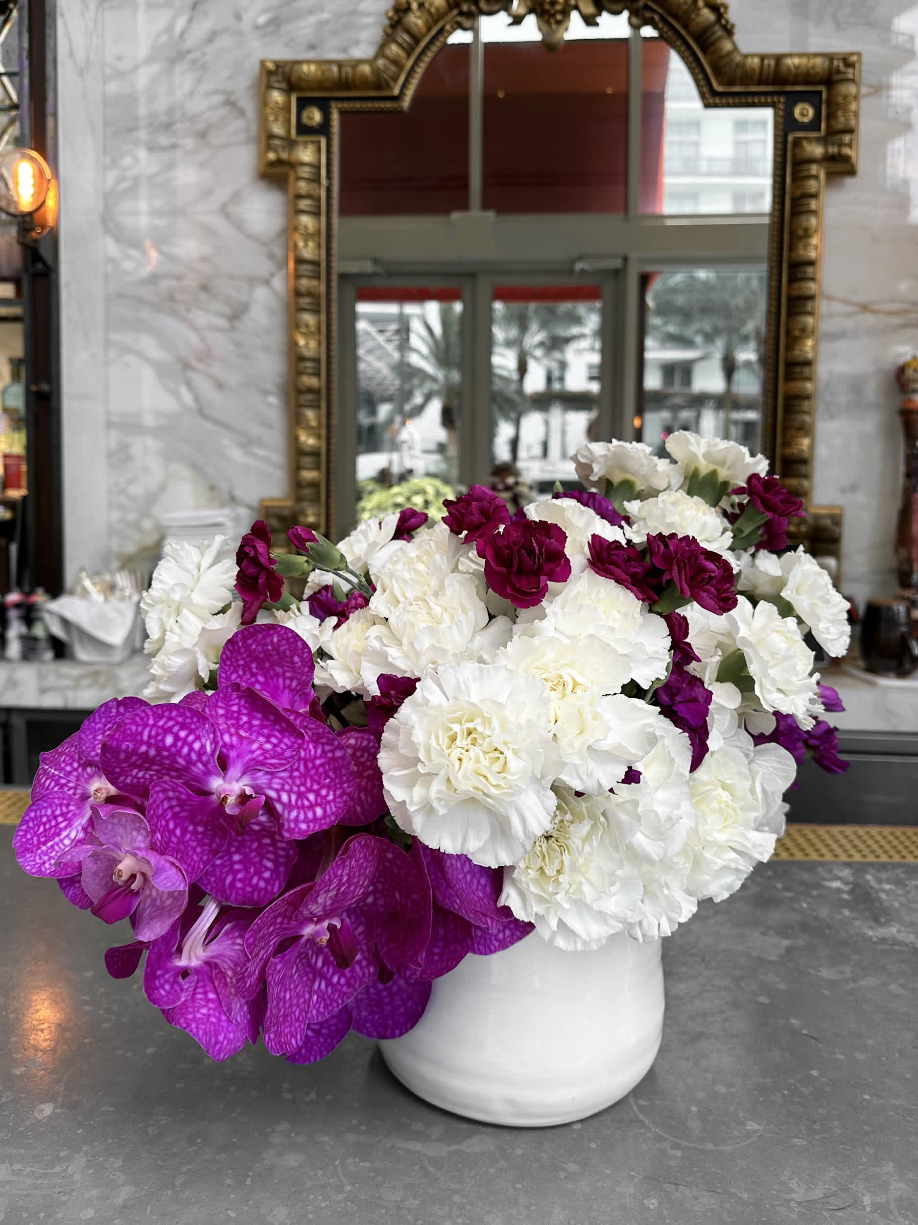 Majestic Orchid Violet Vase - The &quot;Majestic Orchid Violet Vase&quot; is a striking arrangement that combines the elegance of white and deep magenta carnations with the exotic beauty of vibrant purple orchids. This captivating mix is beautifully presented in a sleek white round vase, making it a stunning centerpiece for any occasion. The bold colors and sophisticated design are sure to leave a lasting impression.  Arrangement Details: Main Flowers: White Carnations, Magenta Carnations, Purple Orchids Container: White round ceramic vase Approximate Dimensions: 16&quot;W x 14&quot;H