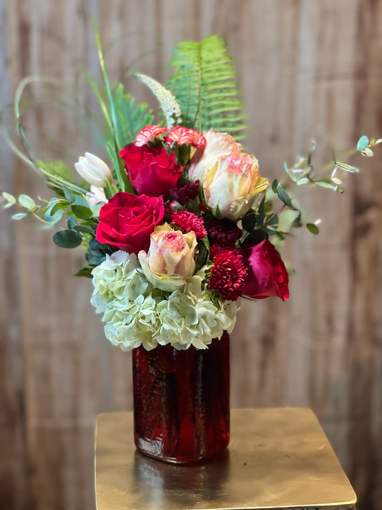 Lavish - The ruby red vase pulsed with vibrant life, overflowing with a symphony of textures and colors. Three velvety red roses, deeply symbolic of love and passion, stood tall and proud, their petals unfurling in graceful curves. Nestled beside them, three blushing pink roses offered a softer, more gentle counterpart, whispering promises of affection and admiration. The airy elegance of a white hydrangea added a touch of ethereal beauty, while clusters of delicate mini carnations created a charming, miniature landscape. Pops of color and playful shapes emerged from the tulips and veronica, adding a whimsical touch. And as a final, heartwarming detail, heart-shaped bear grass playfully danced around the arrangement, sealing the deal with a sweet, visual declaration of love. This wasn't just a bouquet; it was a carefully curated expression of heartfelt emotion, a stunning masterpiece designed to bring joy and warmth to any room.