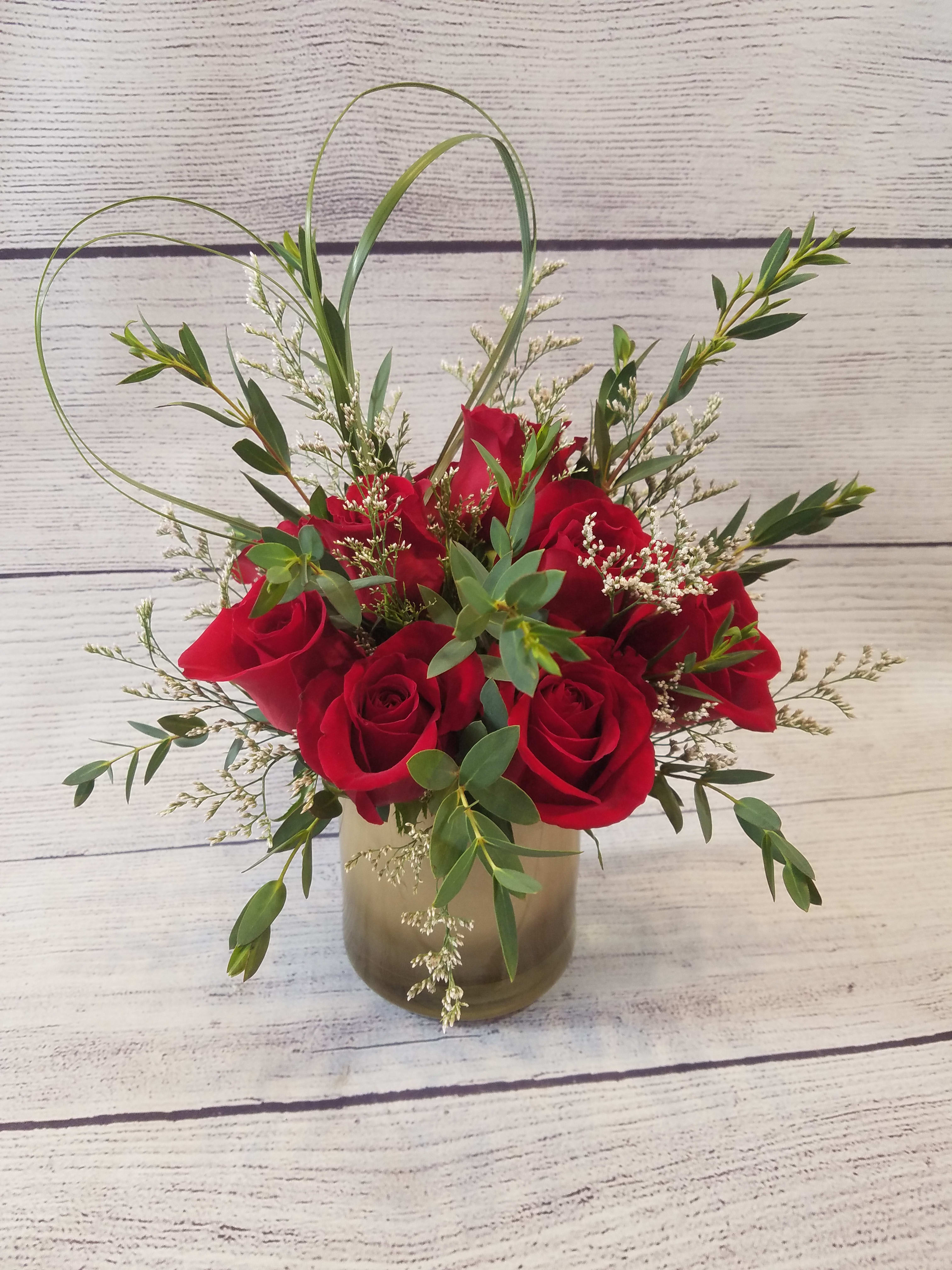 Love is in the Air - This arrangement is more than just a beautiful bouquet; it's a statement of refined affection. The crimson petals of a dozen red roses, symbols of deep love and passion, are perfectly showcased within the smooth, reflective surface of a gold cylinder vase. The addition of fragrant eucalyptus provides a touch of natural elegance and a refreshing scent, while the whimsical bear grass heart injects a playful, heartfelt touch. This stunning display speaks volumes to someone who appreciates a touch of class and sophistication, conveying your feelings in a way that is both timeless and undeniably beautiful. It's the perfect way to say &quot;I love you&quot; with an undeniable touch of grace.