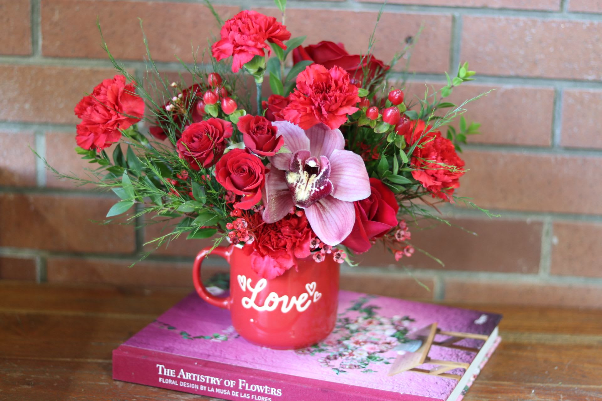 Sweet Sentiments  - &quot;Sweet Sentiments&quot; - A Valentine's Day Gift That Speaks From The Heart  Our charming Valentine's design begins with a cheerful red ceramic mug, featuring the word &quot;Love&quot; in whimsical white script - a heartwarming keepsake that brings joy long after the flowers fade. Inside this special vessel blooms a romantic arrangement that celebrates the season of love.  Each &quot;Sweet Sentiments&quot; arrangement showcases: - A striking red cymbidium orchid as its focal point - Classic red roses for timeless romance - Dainty red spray roses adding delicate charm - Ruffled red carnations providing long-lasting beauty and fullness - Delicate clusters of red wax flowers creating soft texture - Fresh greenery adding natural movement and grace  Perfect for gift-giving, this petite design makes a big impact on a desk, bedside table, or anywhere a touch of love is needed. The premium blooms are carefully selected for their lasting quality, ensuring your sentiment of affection continues to bring joy throughout the Valentine's season.  Height approximately 8-10 inches Available for Valentine's Day delivery February 12-14th Each arrangement is handcrafted - slight variations in flowers maintain the design's romantic character while ensuring the highest quality seasonal blooms.  The perfect choice for someone who appreciates both the beauty of fresh flowers and the practicality of a cherished keepsake mug.​​​​​​​​​​​​​​​​