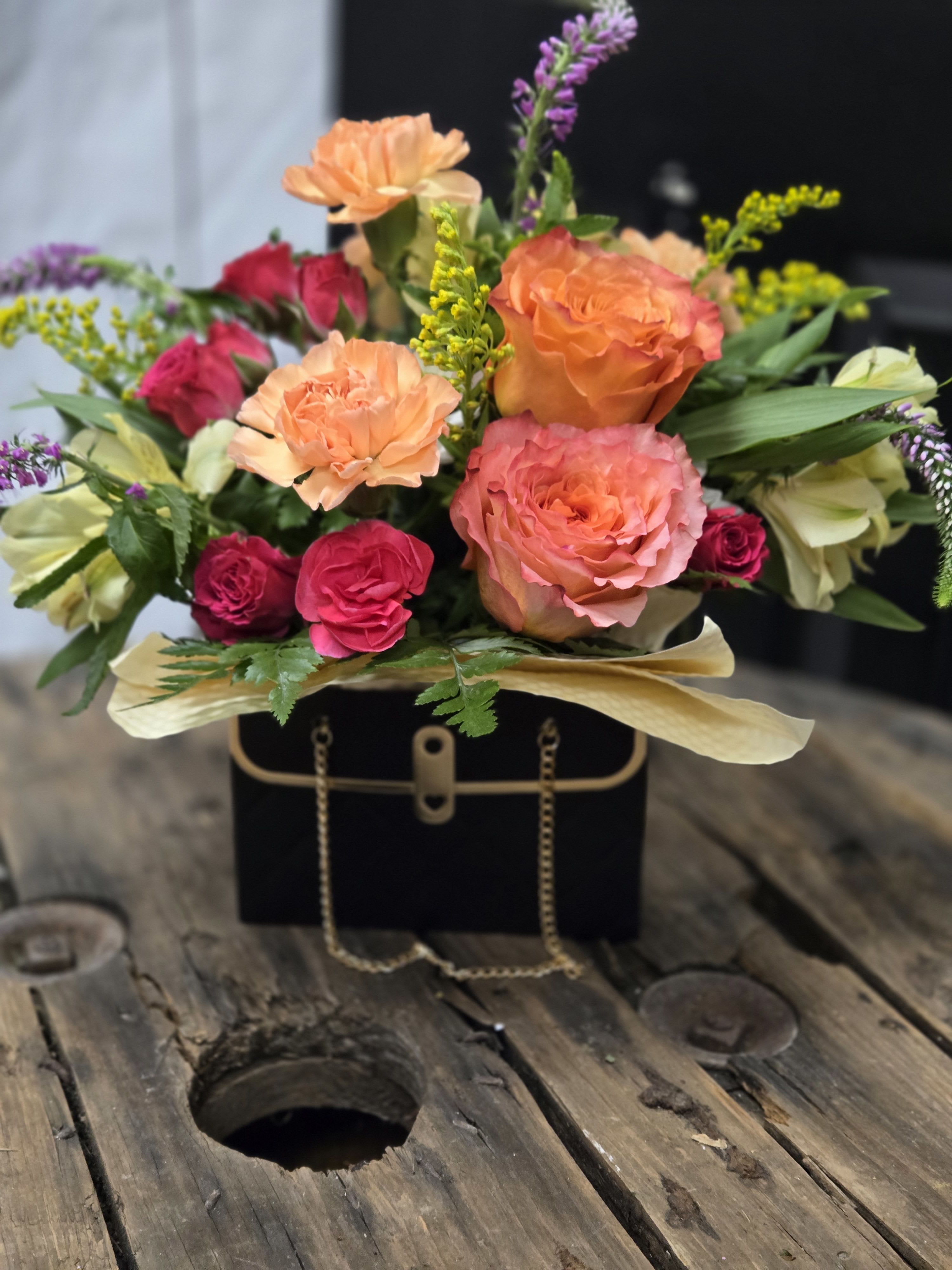Purse Full of Posies  - This floral arrangement is a stunning mix of warm and vibrant colors. It features lush  roses, delicate  carnations, pink spray roses, and soft yellow blooms, all beautifully accented with sprigs of purple flowers and fresh greenery. The bouquet is elegantly presented in a chic black box designed to resemble a stylish handbag ( handbag colors may vary), complete with gold details and a chain handle. The combination of bold hues and sophisticated packaging makes this arrangement both eye-catching and luxurious—a perfect gift for any special occasion!