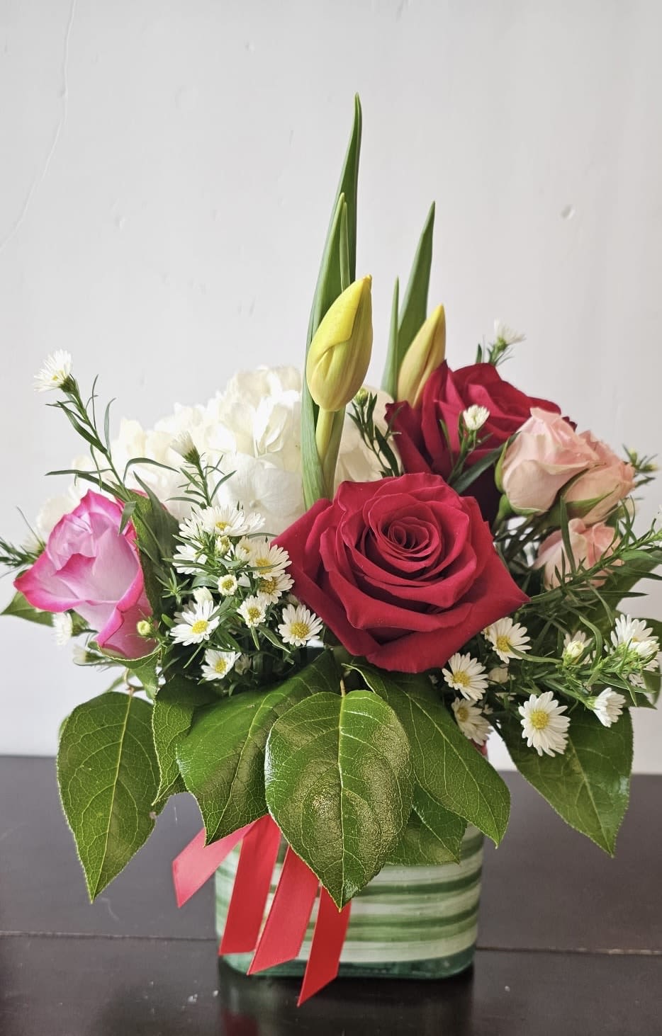 Stacie's Love and Affection - This stunning floral arrangement is a perfect blend of romance and elegance. Featuring lush red and pink roses, delicate white hydrangeas, and cheerful daisy-like blooms, it is beautifully accented with fresh greenery. The arrangement is complemented by graceful yellow tulip buds, adding a touch of sophistication. Presented in a chic striped green vase and tied with a charming red ribbon, this bouquet is ideal for any special occasion, from anniversaries to heartfelt celebrations.  Perfect for gifting or as a centerpiece, this arrangement brings warmth, beauty, and timeless charm to any space.