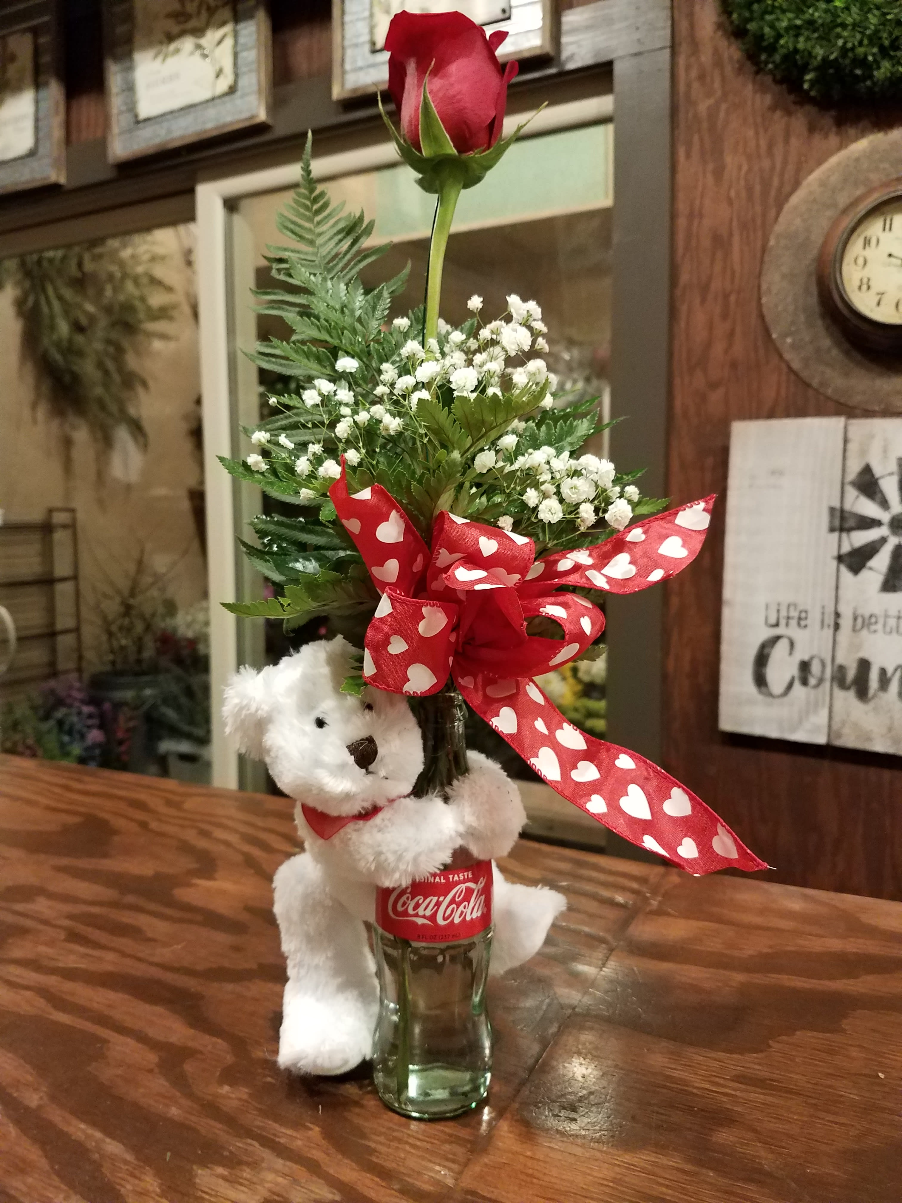 A Coke and a Smile  - This design features a plush teddy bear which has arms attached. The bear placed onto the bottle which hold a single rose with greenery , babies breath and a bow.