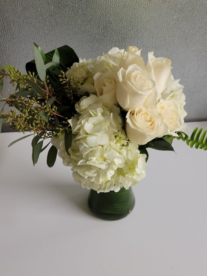 White Simplicity - White Hydrangea, white roses uniquely designed in a 8 inch gathering vase, accented with flat fern, seeded eucalyptus and aspidistra leaves.