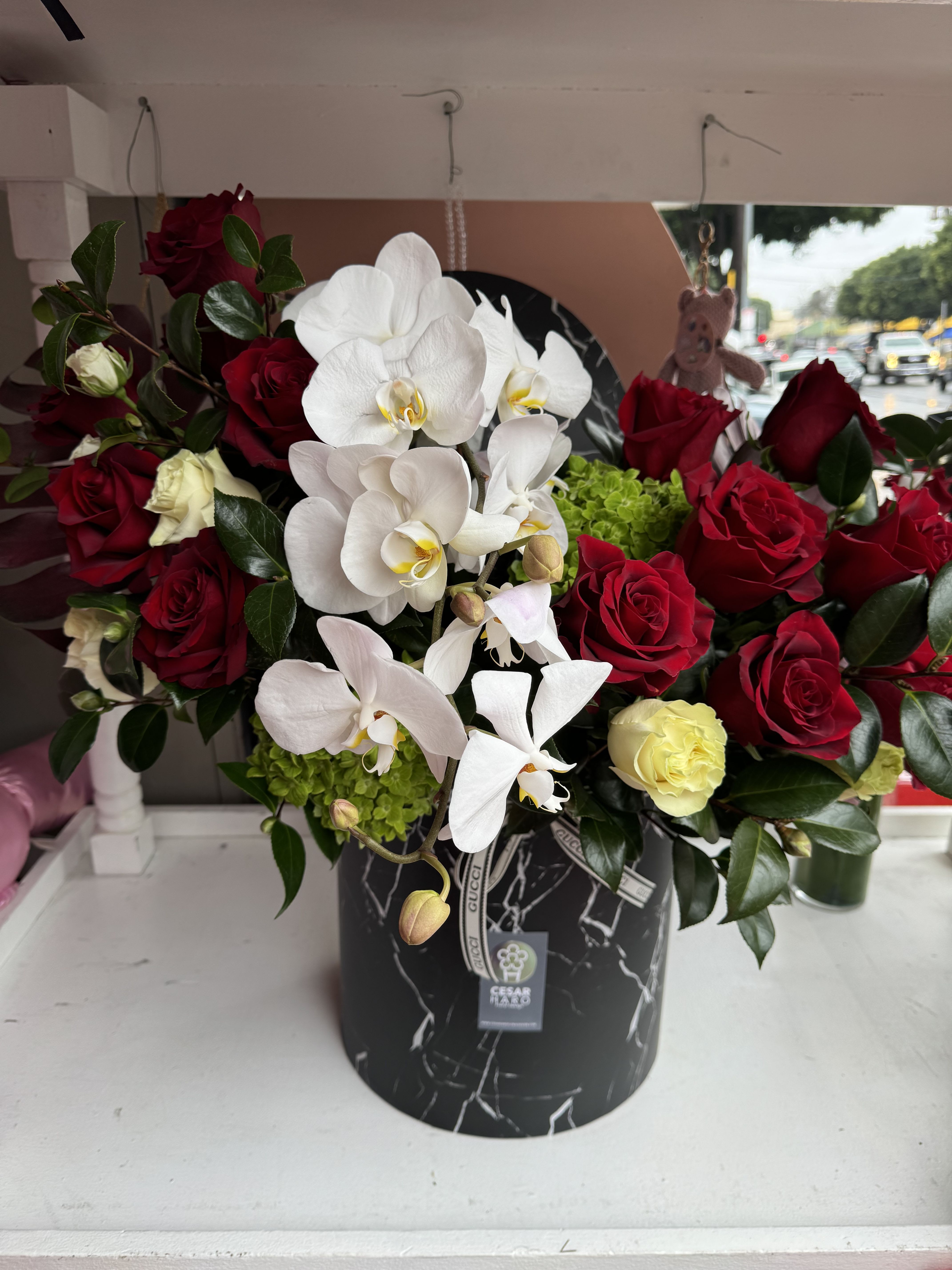 Designed for Elegance - A sophisticated arrangement set on a sleek faux black marble base, featuring pristine white orchids, hydrangeas, alongside luxurious red and cream roses. The designer touches elevate this refined display, blending timeless beauty with modern grandeur for a truly elegant composition.