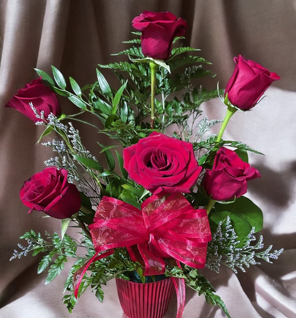 Red Bliss  - This elegant arrangement features six vibrant red roses as the centerpiece, nestled among lush greenery and delicate filler flowers. The roses are beautifully accented by ferns and sprigs of soft, silvery foliage, adding texture and depth. A bold red ribbon bow ties the arrangement together, creating a striking yet balanced display.