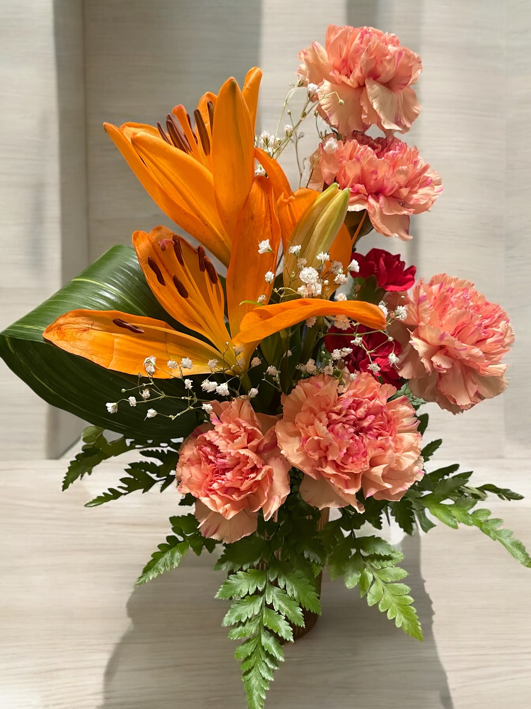 Sunset Charm  - This vibrant arrangement radiates warmth and charm, featuring bold orange lilies as the focal point, complemented by soft peach carnations that add a gentle, romantic touch. Delicate sprigs of white baby’s breath provide an airy contrast, while pops of deep red blooms add richness and depth.  Note*: In some cases the lilies in this design might come in white instead of orange, due to seasonality and availability. 