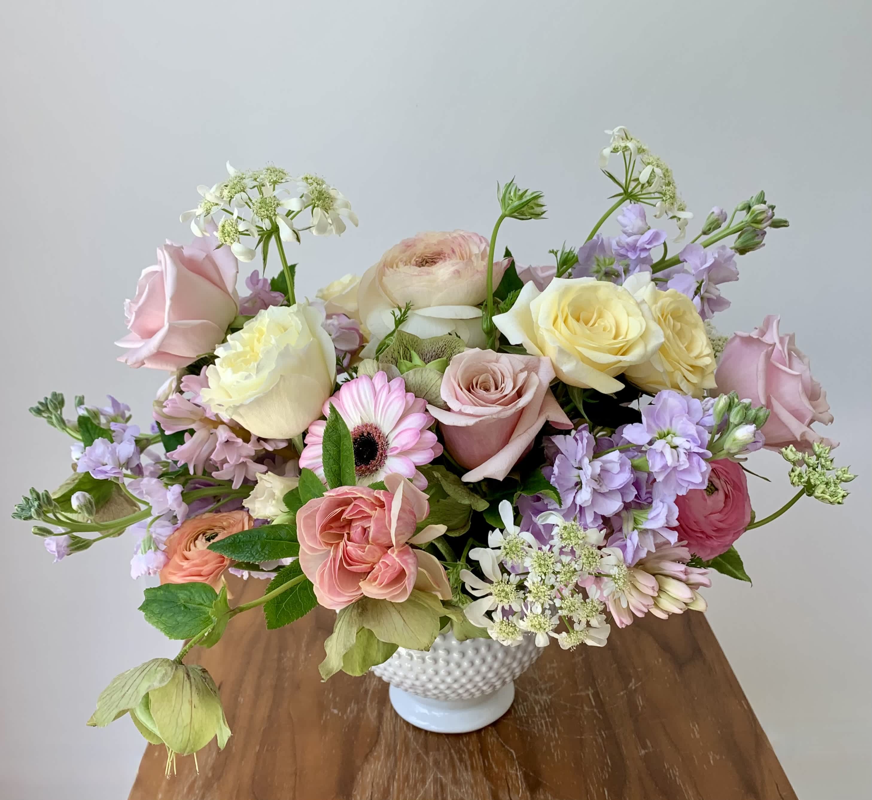 Serena  - Fresh flower centerpiece with soft color blooms arranged in a small ceramic compote vase. The focal flowers are premium roses, germinis, ranunculus, stock, lisianthus and complementing fillers and greens to create a lovely garden style arrangement. 
