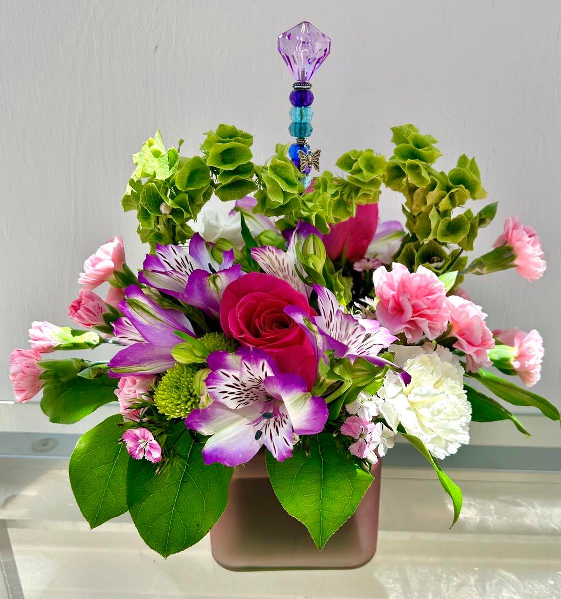 Mi Amor - Heart shaped Flower in a spring colored vase with spring flowers to match. Color of vase could be Pink, Purple, Yellow, or Green. Heart shaped flowers could be made of, larkspur, bells of Ireland, or babies breath. We will pick the freshest option and make it beautiful.  Also comes with a wand with a charm on it, the wands range in color.