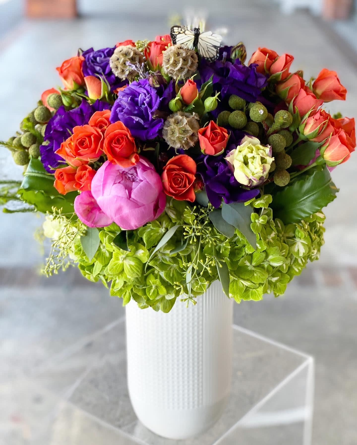 Madison - Orange roses and Purple and green flowers  - Tall and elegant! In our new white ceramic vessels, this abundant design is made of orange, purple, and green hues - roses, lisianthus, hydrangeas, peonies, and more.