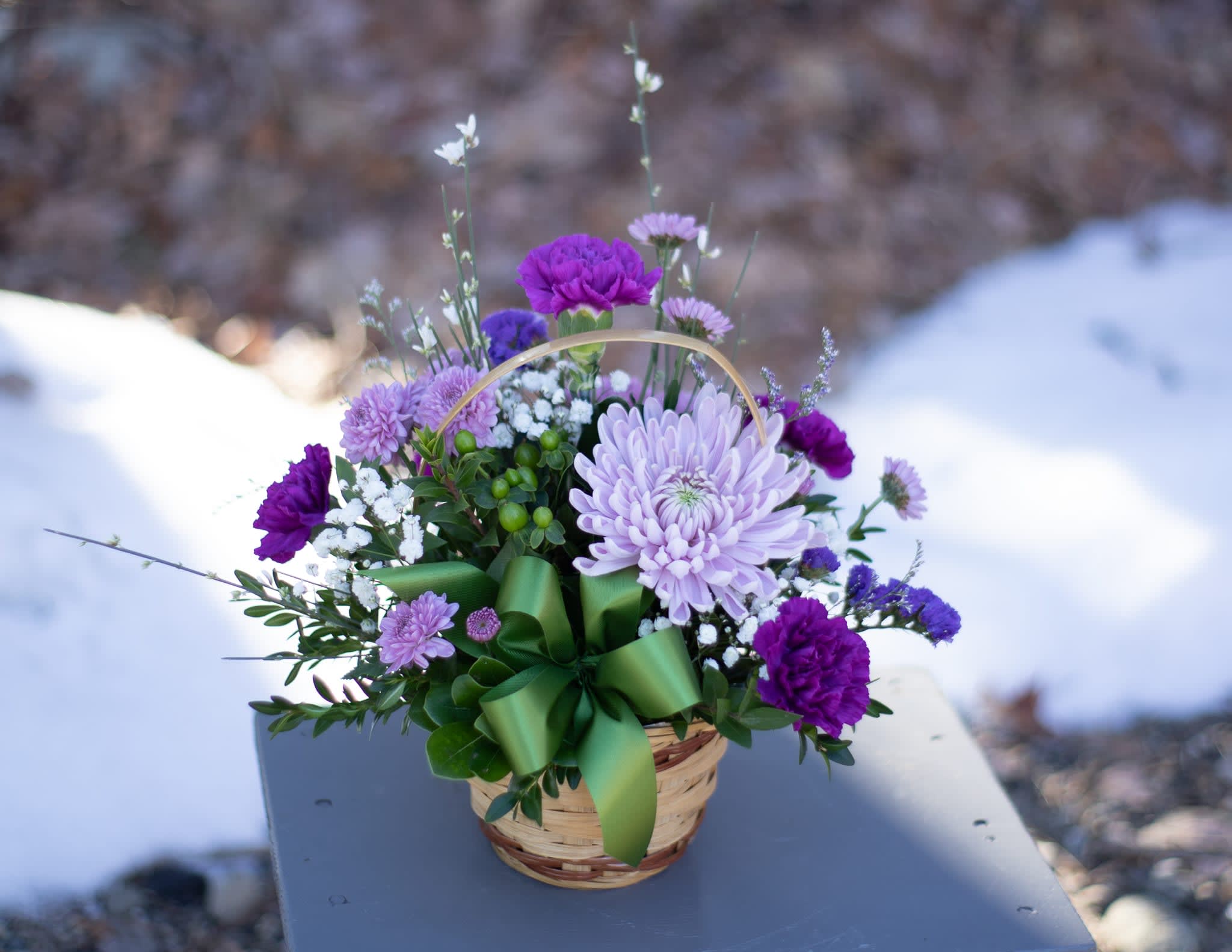 Grape Expectations Basket - Complimentary color blooms with a twist. Standard blooms in a wicker basket. Arranged in traditional all around fashion in a 5 inch basket. Purple carnations, lavender cremons, green hypericum berries with splashes or daisy, button or cushion poms. Upgrades will include more premium grade flowers like hydrangeas and roses. 2024 Baskets have no handle