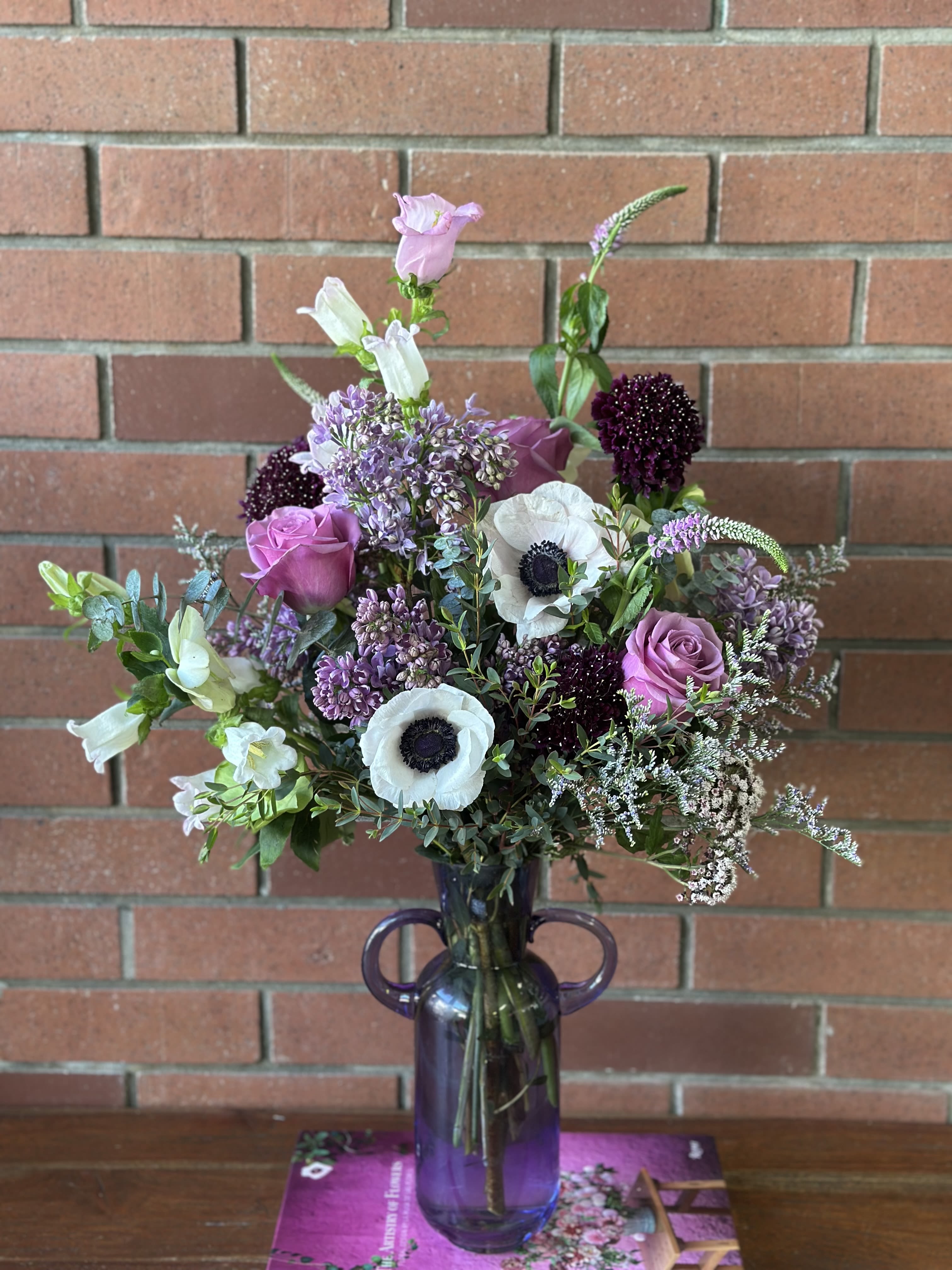 Twilight Garden’s Dance  - Here's a description that highlights the fresh lilacs while acknowledging all the special elements of this romantic arrangement:  &quot;'Twilight Garden's Dance' celebrates the fleeting beauty of fresh-cut lilacs in their peak season. These fragrant purple lilac blooms are thoughtfully paired with garden-inspired flowers including striking white anemones, mauve roses, and deep burgundy scabiosa. Delicate white bell flowers and airy astilbe add movement and grace, while fresh eucalyptus and greenery provide a lush foundation.  This seasonal arrangement showcases the natural beauty and subtle fragrance of spring lilacs, expertly combined with premium blooms to create a romantic, garden-gathered style. Presented in our signature purple glass vase, this design captures the enchanting moment when twilight touches a spring garden.  Note: Fresh lilacs are a seasonal treasure, available for a limited time during peak spring bloom. Their delicate nature and short vase life make them an extra special addition to your bouquet. To maximize enjoyment: - Keep away from direct sunlight and heat - Change water daily - Display in a cool location - Expect lilacs to last 3-5 days, while other blooms will continue their display