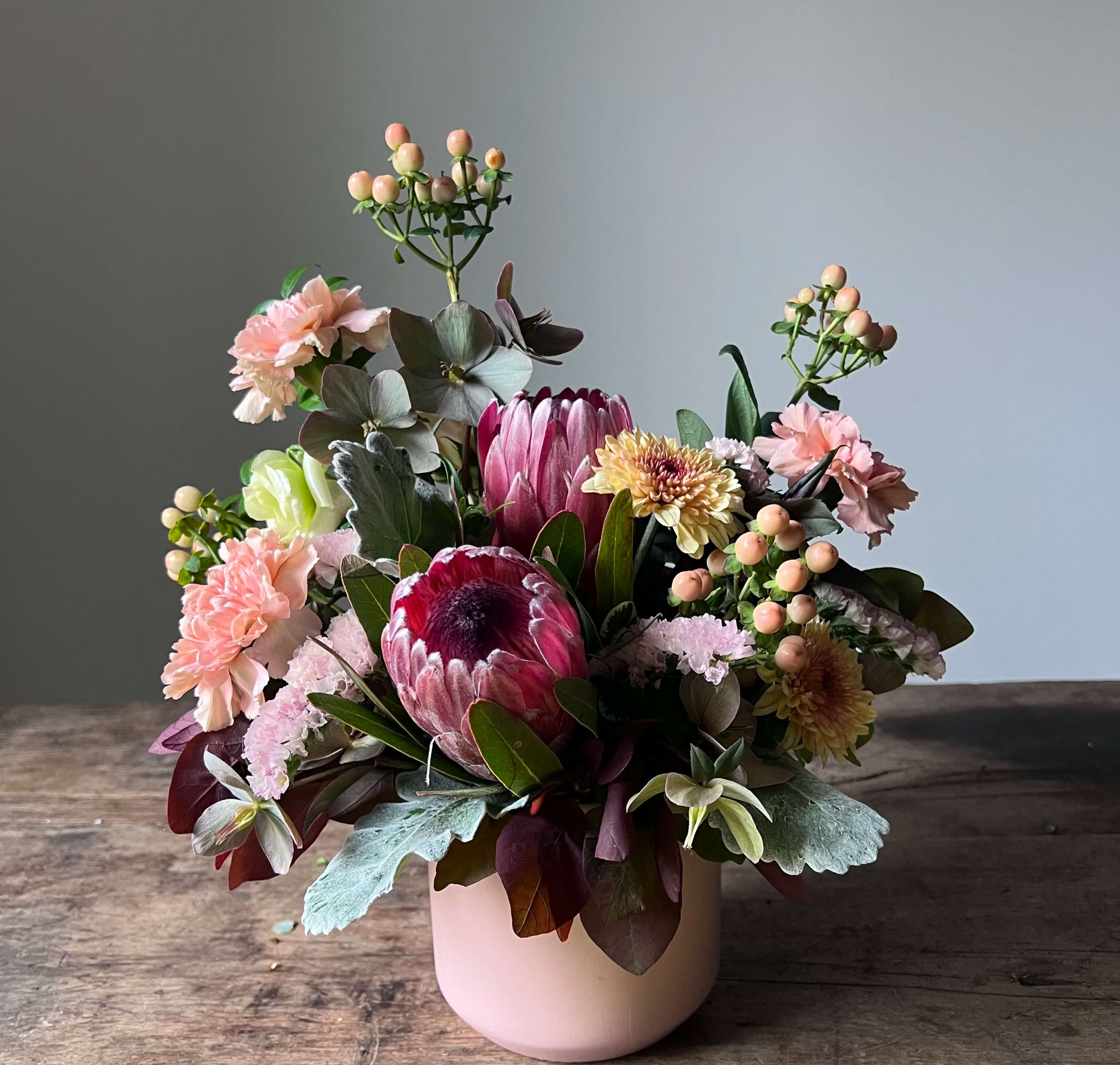 Exotica Exotica - This moody gathering of eclectic blooms is a sophisticated palette of burgundy, peach, silver and muted golden tones. Blooms include protea, hypericum, antique carnations and more. Ceramic vase.