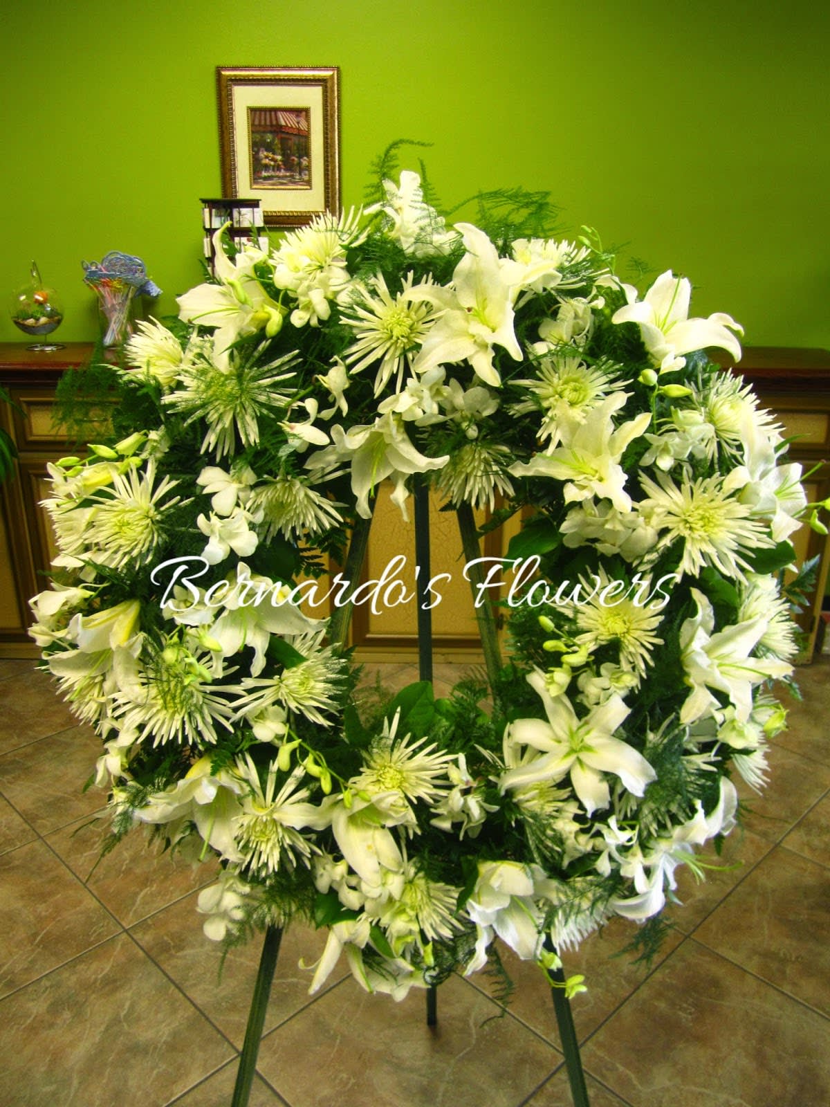 BFS-W16 Sympathy Wreath - Our Sympathy White Wreath is a heartfelt expression of condolences and reverence. The combination of white lilies, mums, and Dendrobium orchids creates a serene and tranquil arrangement, offering comfort and support during a difficult time. Whether displayed at a funeral service or sent as a gesture of sympathy, this wreath serves as a lasting symbol of peace and solace for those mourning the loss of a loved one. 26 Inches in Diameter