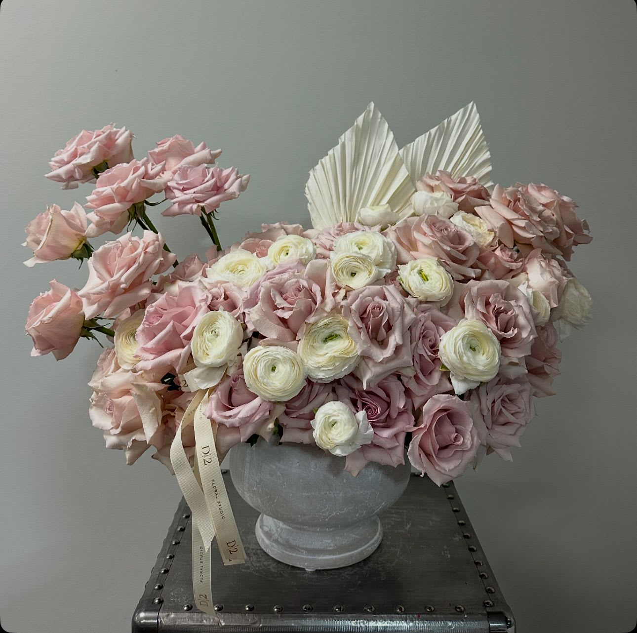 Josephine Josephine - This floral arrangement features a luxurious blend of soft pink and blush roses, complemented by delicate white ranunculus. The flowers are artfully arranged in a classic white vase, with two white dried palm leaves adding a modern, sculptural touch. A subtle ribbon with elegant branding cascades from the arrangement, enhancing its sophisticated and romantic aesthetic. The overall design exudes elegance and timeless beauty, making it perfect for special occasions or upscale decor.