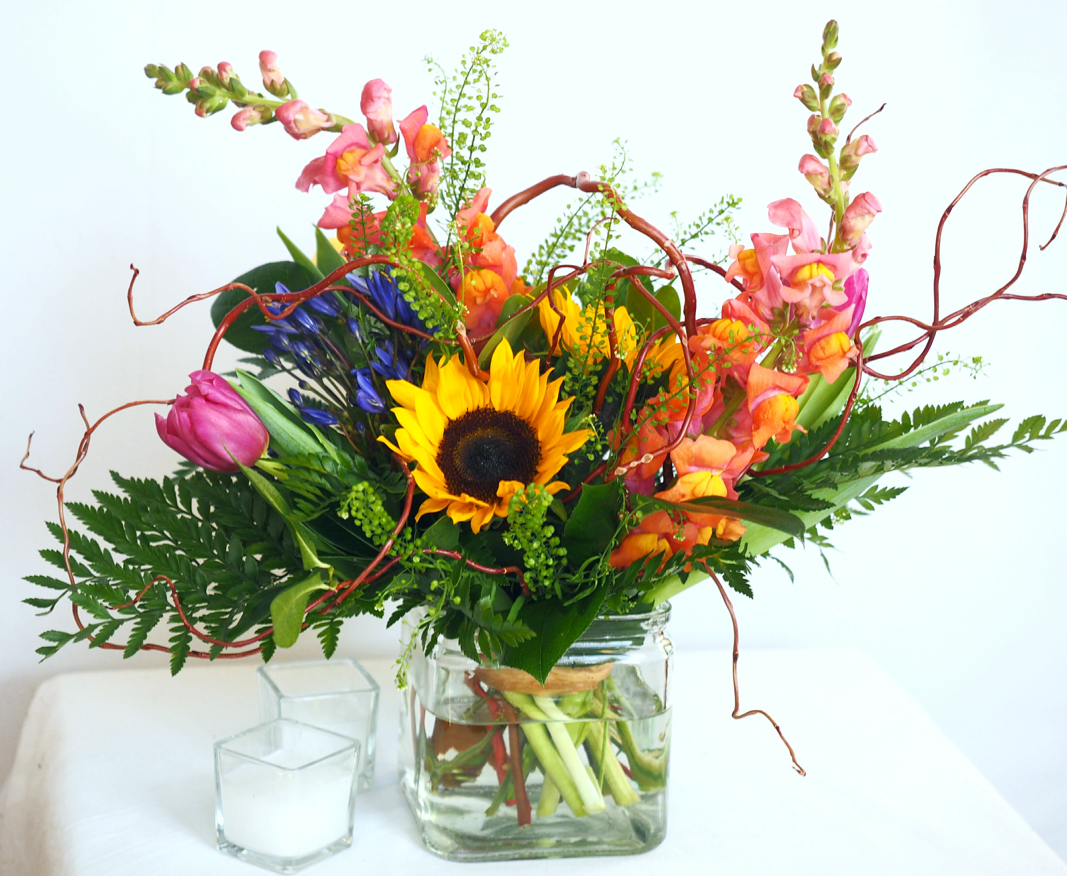 Dorothy - Colorful. Uplifting. Whimsical.   This cheerful, garden-style display features sunflowers, snapdragons or stock, and fresh tulips designed in a clear glass rectangular vase.   Please note: During the winter season, sunflowers can be a challenge in terms of quality and longevity. We will always do our best to match the photo- however, we will occasionally need to make a substitution.  *SYMPATHY/FUNERAL: PLEASE NOTE THE TIME, PLACE, &amp; NAME OF THE DECEASED FOR THE SERVICE IN THE SPECIAL NOTES SECTION OF YOUR ORDER - WE ARE UNABLE TO GUARANTEE DELIVERY TIME WITHOUT THIS.*