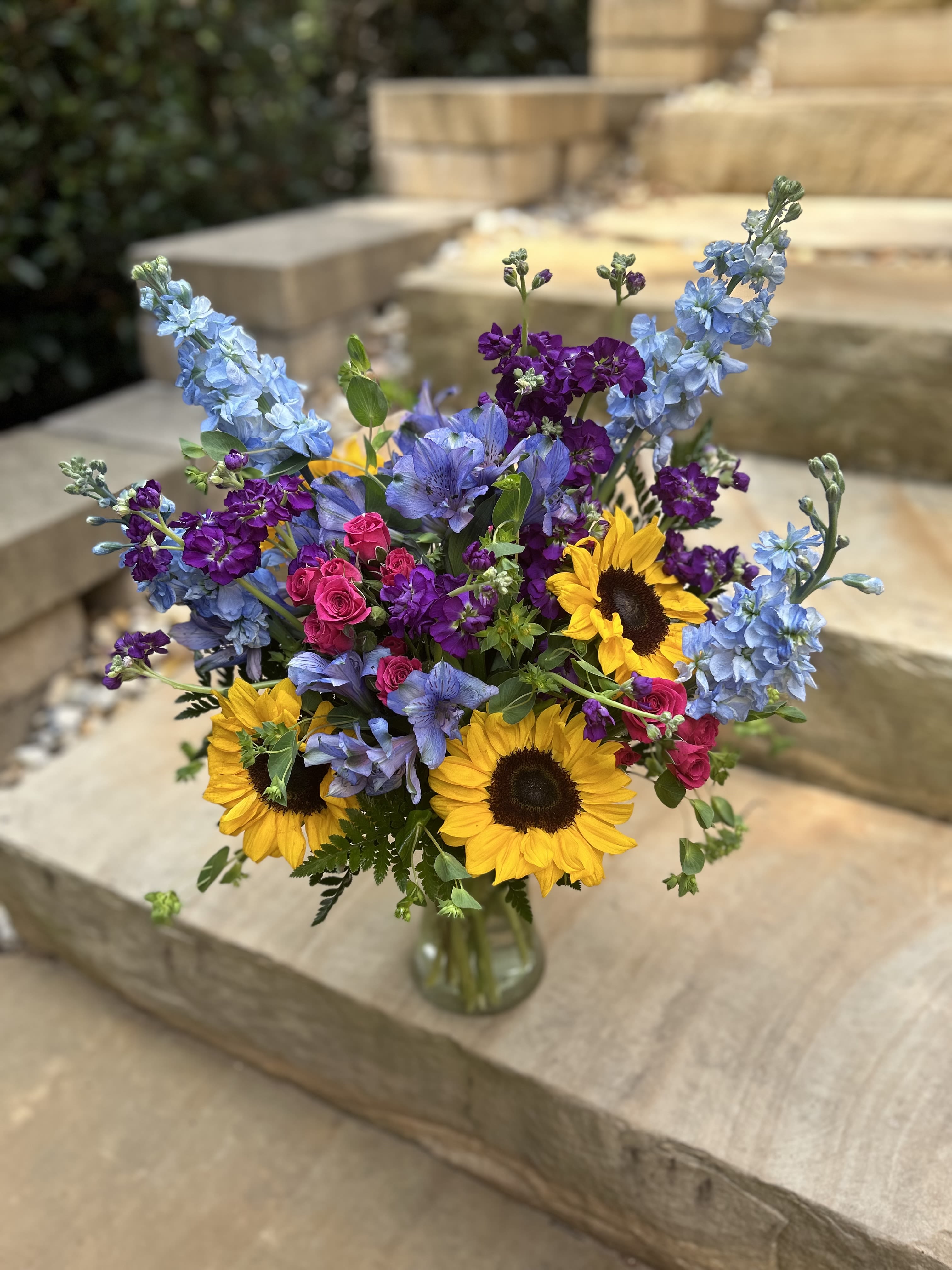 Sunny &amp; Blue - Sunflowers, stock, alstroemeria, spray roses and greenery in shades of blue, purple, hot pink and yellow in a tall glass vase. 