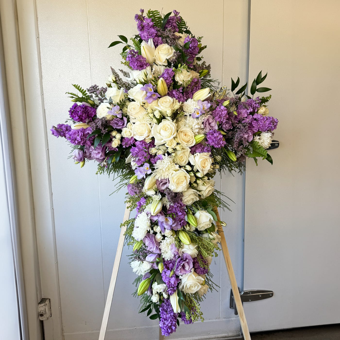 PURPLE N WHITE CROSS - purple, pink, white flowers, and greenery. The standing cross includes the wood easel. Pink roses, white hydrangeas, spray roses, orchids, seasonal florals, and greenery