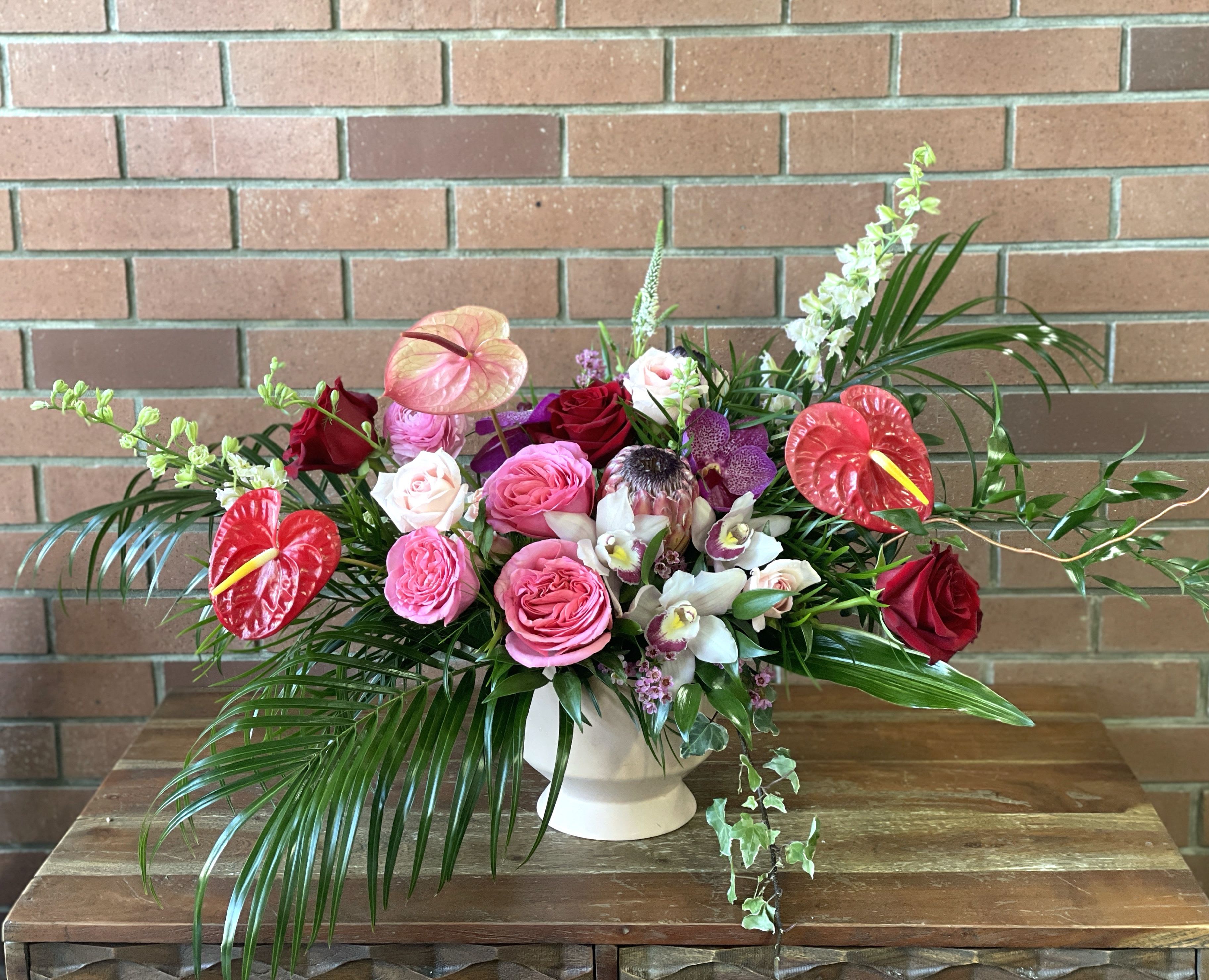 Tropical Garden Romance  - &quot;Tropical Garden Romance artfully marries bold tropical elements with soft garden blooms in a luxurious modern design. Showcased in our contemporary pink ceramic vase, this striking arrangement features dramatic red anthurium blooms and lush palm fronds that create an exotic foundation. Premium garden roses in shades of blush pink and deep burgundy provide romantic softness, while elegant white orchids add graceful exotic beauty.  The design is thoughtfully layered with delicate pink ranunculus and vertical blooming stems that create enchanting height and movement. Fresh palm fronds sweep dramatically outward, while trailing greenery adds an organic finishing touch to this sophisticated tropical-garden fusion.  Height: Approximately 24-26&quot; including vase Width: Approximately 28-30&quot; Style: Modern Tropical-Garden Fusion Care: Fresh water daily; avoid direct sunlight Longevity: 7-10 days with proper care  Perfect for: - Luxury homes - Special celebrations - Hotel lobbies - Corporate spaces - Intimate events  Each Tropical Garden Romance arrangement is handcrafted to order, ensuring a unique statement piece that brings together the best of tropical drama and garden elegance.&quot; 