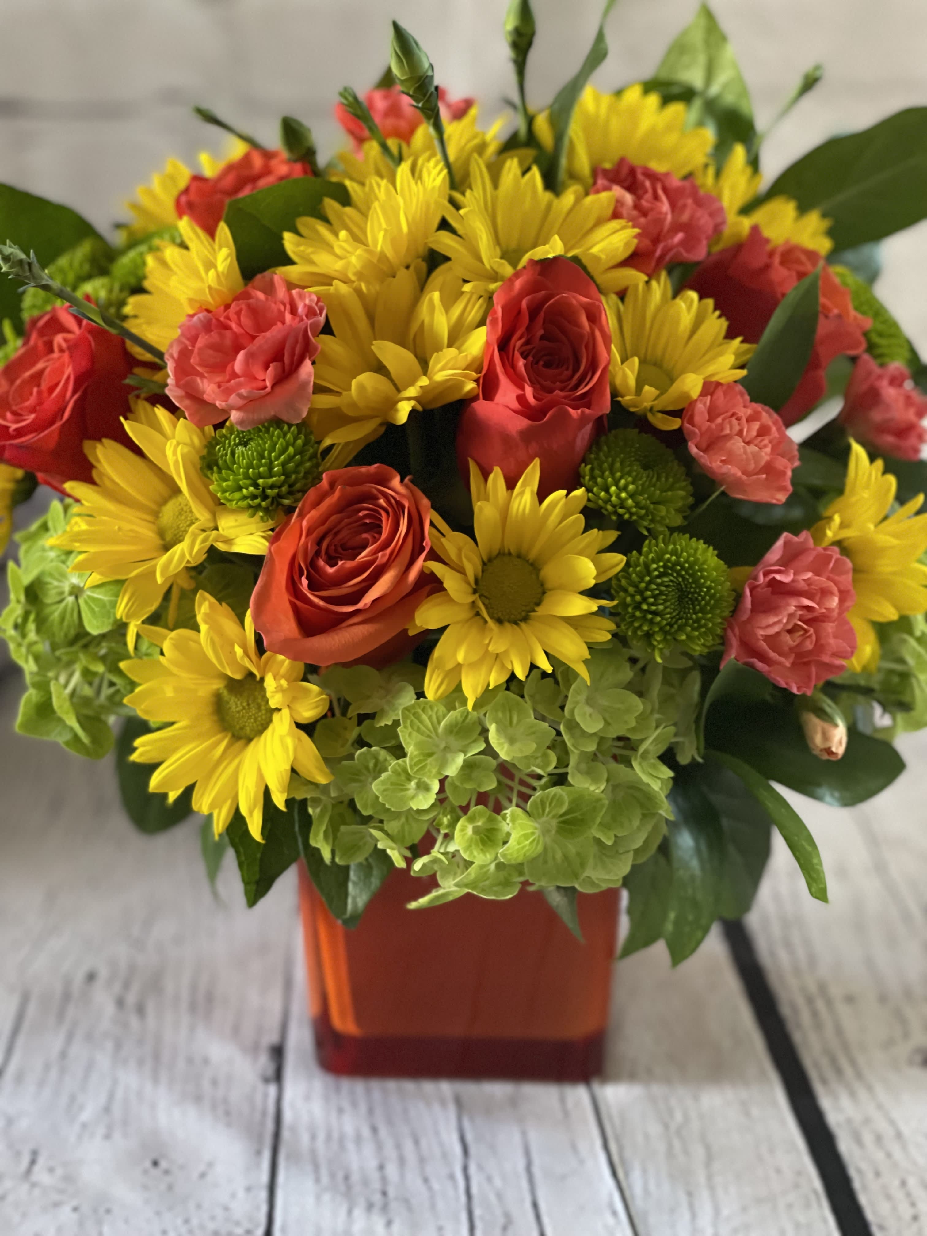 Citrus Zest Spring - Our Citrus Zest Spring floral arrangement is a bright and cheerful bouquet celebrating the season's arrival. This stunning arrangement features a perfect blend of roses, hydrangeas, and daisies in a gorgeous orange glass cube vase.  The orange roses are a symbol of passion, energy, and excitement, and they complement the green hydrangeas perfectly, representing forgiveness, freshness, and renewal. The yellow daisies symbolize happiness, joy, and new beginnings, and are known for their ability to bring sunshine to even the darkest of days. Together, these blossoms create a harmonious union of colors, fragrances, and emotions.  The Citrus Zest Spring floral arrangement is a perfect choice for celebrating spring, ceremonies, birthdays, or as a thoughtful gift for the ones you love. This radiant and lively bouquet will brighten anyone's mood or home, reminding them of the joy and happiness that the season brings.  Order the Citrus Zest Spring floral arrangement today, and let our beautiful bouquet add a touch of warmth, sweetness, and positivity to your life or the person you care for.