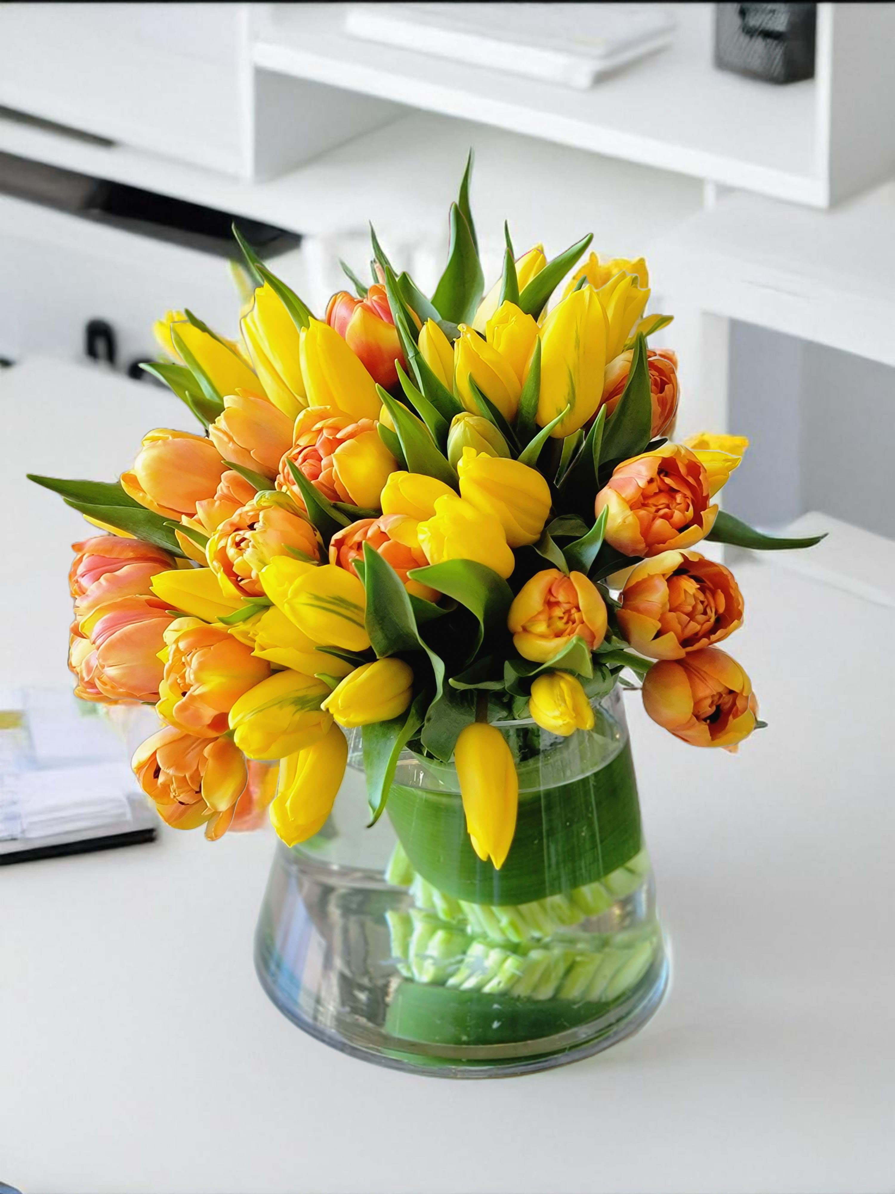 Sunny Tulips  - This luxurious tulip arrangement features vibrant yellow and orange tulips, artfully designed in a modern glass vase. Perfect for brightening any office, or special occasion, this stunning floral piece is a timeless gift that radiates warmth and joy.