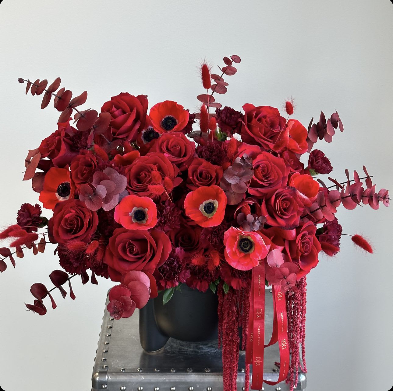 Ruby Ruby - This is a luxurious floral arrangement in a bold monochromatic red palette, featuring deep red roses, vibrant red anemones with dark centers, rich burgundy carnations, and striking red-tinted eucalyptus. The arrangement is enhanced with unique textural elements like hanging amaranthus and fluffy red bunny tails, giving it a dramatic and opulent look. Presented in a sleek black vase and adorned with elegant red ribbon, this design exudes sophistication and passion.