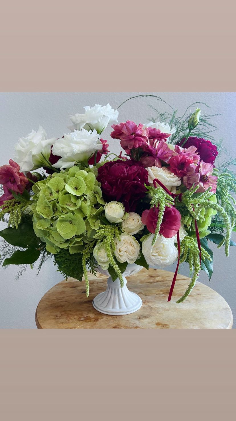 Winter Elegance Winter Elegance - This is April May Flowers' unique vision of winter elegance. This floral sculpture features a harmonious blend of seasonal blooms and greenery, designed to capture the spirit of the holiday season. At its core, rich red peonies and butterfly ranunculus alongside pristine white lisianthus and roses are used, symbolizing love and purity, respectively. These flowers are complemented by lush holiday branches, such as pine or fir, which add a fragrant, wintry touch. Accents of deep greens, adorned with bright reds, introduce a classic Christmas vibe, while delicate accents, like ornamental twigs or ribbon, provide a touch of sophistication. To enhance the arrangement, you will find hanging amaranthus peeking through the greenery. The arrangement is typically presented in an elegant compote dish which adds to its festive charm. Whether used as a centerpiece for holiday gatherings or a lovely gift, this elegant Christmas floral arrangement brings warmth and beauty to any setting, embodying the joy and celebration of the season.
