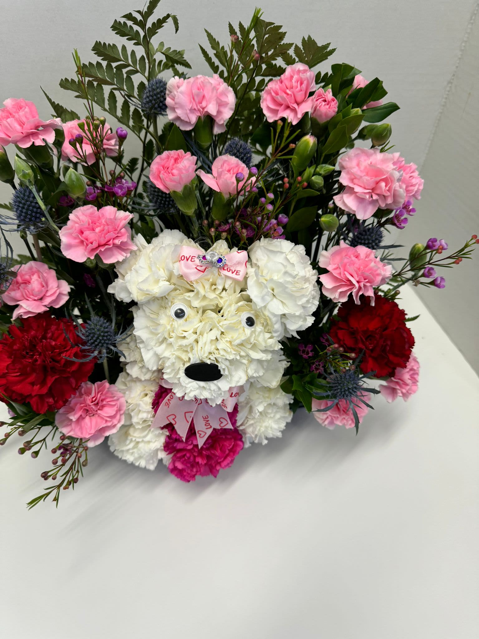 Puppy Flower Arrangement  -  Talk about puppy love! Crafted from lasting white carnations, our canine cutie , surrounded by a mix of blooms beautiful. The perfect pick for the persons they love puppys. Please select the color you want the arrangement to be.