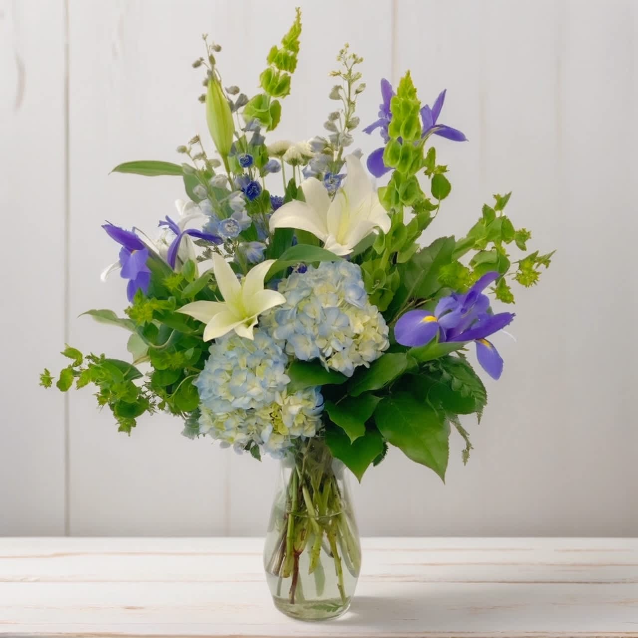 Blue Skies Above - Feeling inspired by the beauty of nature today? Hignight’s has the perfect thing—our stunning Blue Skies Above bouquet! This delightful arrangement features a blend of blue hydrangeas and iris, white lilies, and green Bells of Ireland.  Let these beautiful blooms transport you to a serene garden, where clear blue skies stretch endlessly above. The subdued blue hues of the hydrangeas and bright blue iris, along with the pure white lilies, create a calming and uplifting atmosphere.  Embrace the tranquility and elegance that our Blue Skies Above bouquet brings, and let it inspire your loved ones to find beauty in everyday moments. Whether it's a simple walk in nature or a quiet moment of reflection, these flowers serve as a reminder to appreciate the magic that surrounds us.  Share this bouquet with someone who appreciates the beauty of nature! Let's spread the joy and positivity that flowers bring.  Hignight Florist delivers flower arrangements, wedding flowers, funeral flowers, plants, and gifts in the Quad Cities: East Moline, Moline, Rock Island, Milan, Coal Valley, Silvis, Carbon Cliff, Hampton, Colona, Bettendorf, and Davenport. 