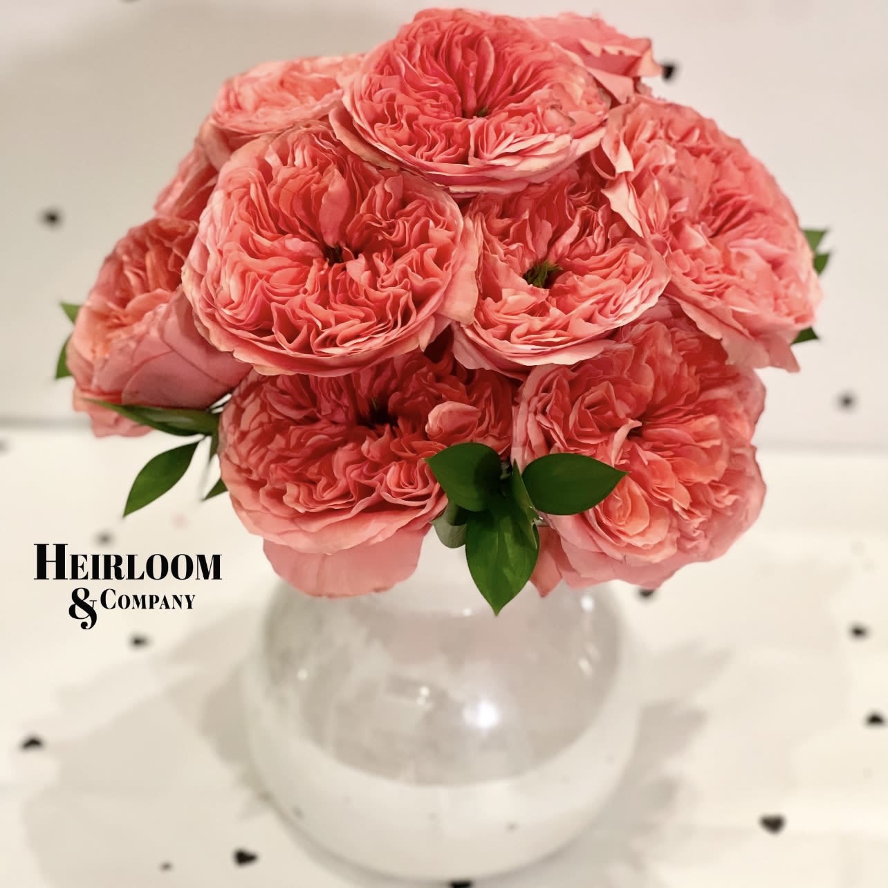 Strawberry Fields  - A breathtaking arrangement of lush, ruffled garden roses in a warm coral hue, thoughtfully designed to bring timeless beauty to any space. Each bloom is carefully selected for its intricate petal structure and rich fragrance, creating a romantic and elegant display. Accented with fresh greenery and arranged in a modern white vase, this bouquet embodies sophistication and charm.  Perfect for special celebrations, or as a luxurious gift, our Garden Rose Elegance bouquet is a signature piece from Heirloom &amp; Company—where every floral creation tells a story.