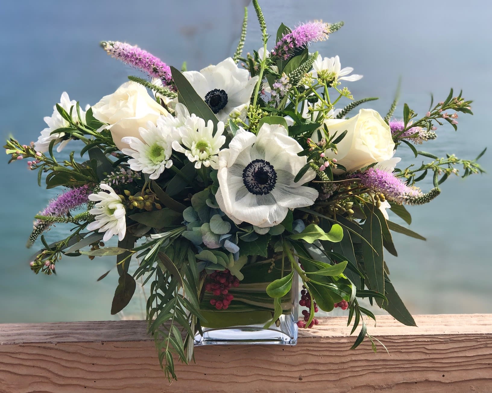 Abalone Cove - A collection of white flowers with accents of blue and lavender tones in a clear glass cube lined with variegated leaf.  Flowers might include hydrangea, veronica, roses, snapdragons, anemones, herbs and textures.
