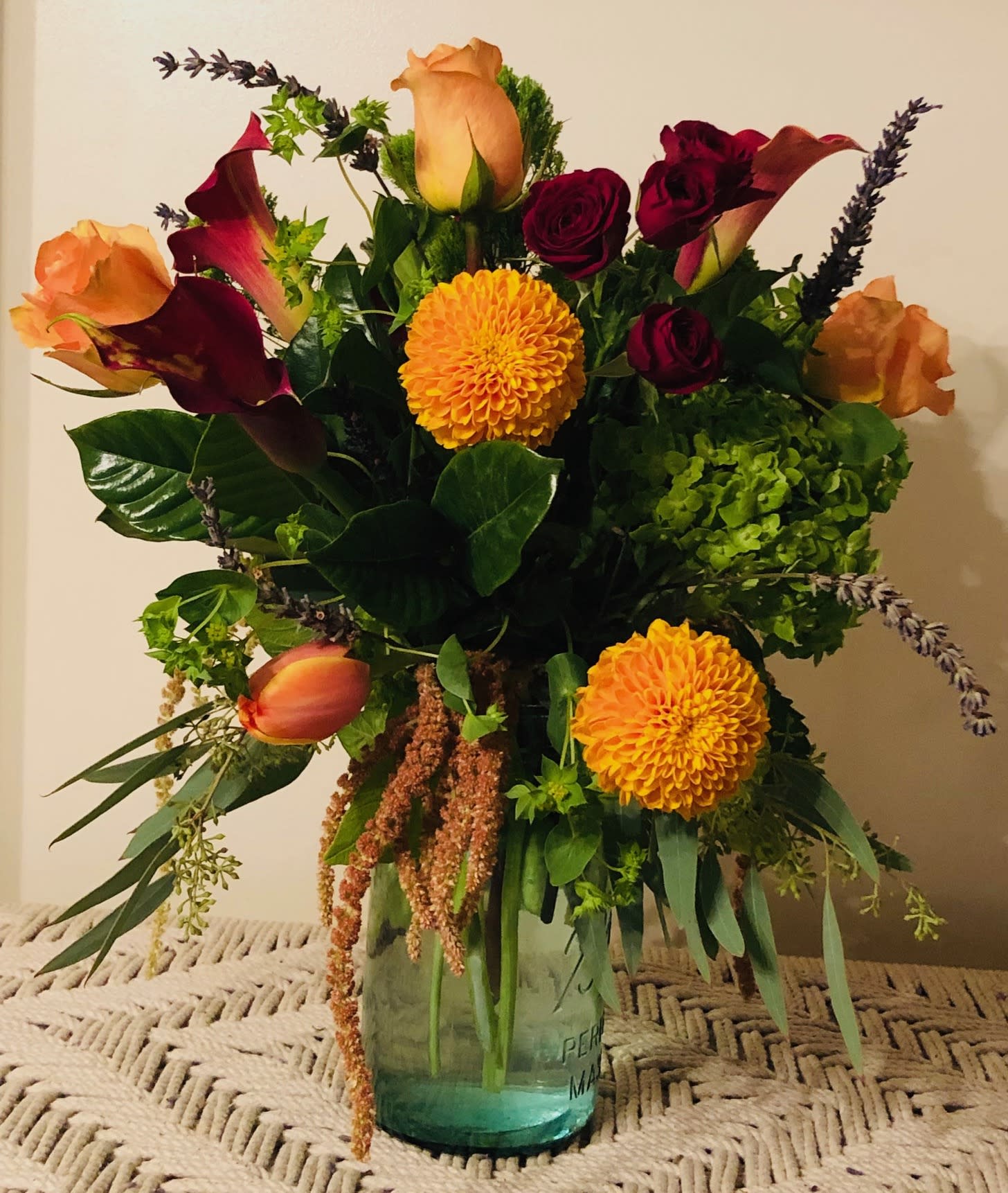 Fab Fall - A rich, jewel colored arrangement of burgundies paired with orange, tangerine, vibrant green hydrangea and a hint of purple/blue tones.  All set off with seasonal textures such as dianthus, eucalyptus and velvety amaranthus in a vintage aquamarine glass Maison jar vase.