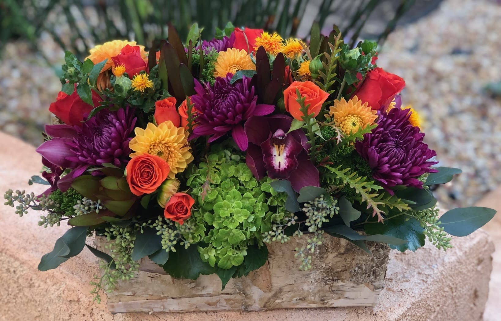 Fall in Birch - Vibrant fall blooms in an oblong birch box. Featuring Dahlias, Roses, Orchid blossoms and lush autumnal textures such as seeded Eucalyptus .  Perfect t as a gift or centerpiece for you Fall festivities.