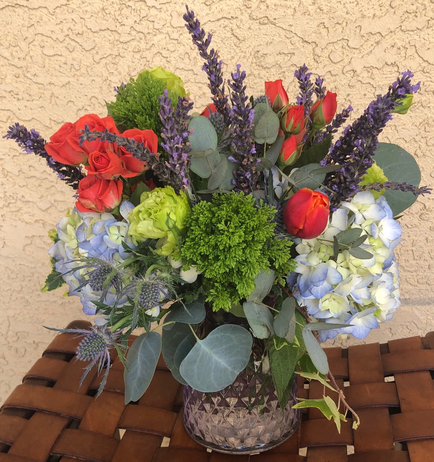Camille - Stunning arrangement of Blue Hydrangea, Vermillion Red Spray Roses and Tulips, Fresh Lavender, Thistle and Green Lisianthus.  All beautifully accented with Eucalyptus, Ivy and other seasonally fresh textures in a  vintage, amethyst cut vase. 