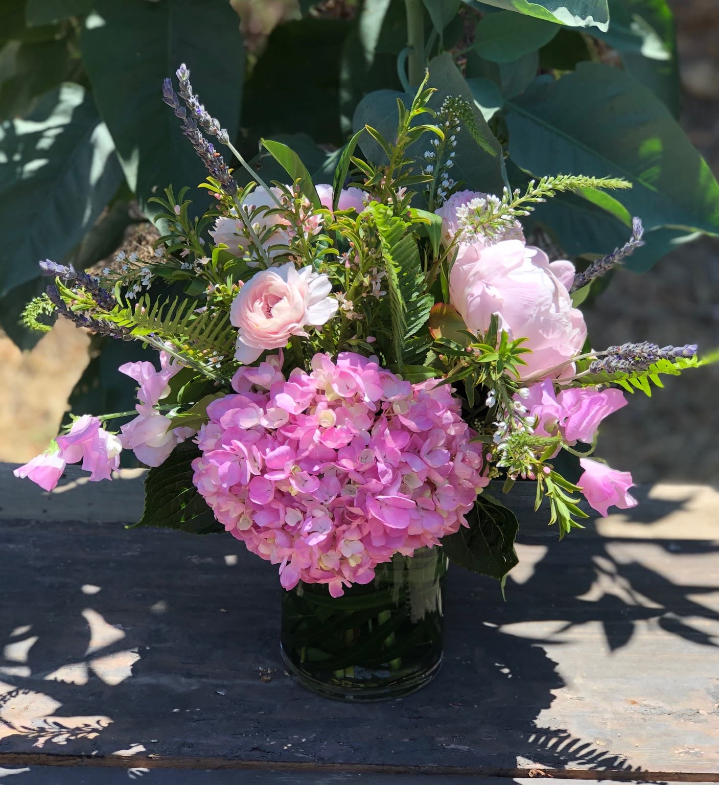 Spring Offering - A collection of mixed pink blooms with a lavender accent and fresh green texture in a tall clear glass cylinder.  Flowers may include hydrangea, Peony, roses, ranunculus, cymbidium orchids curly willow and other seasonal flowers in this color pallet that are fresh from that day.