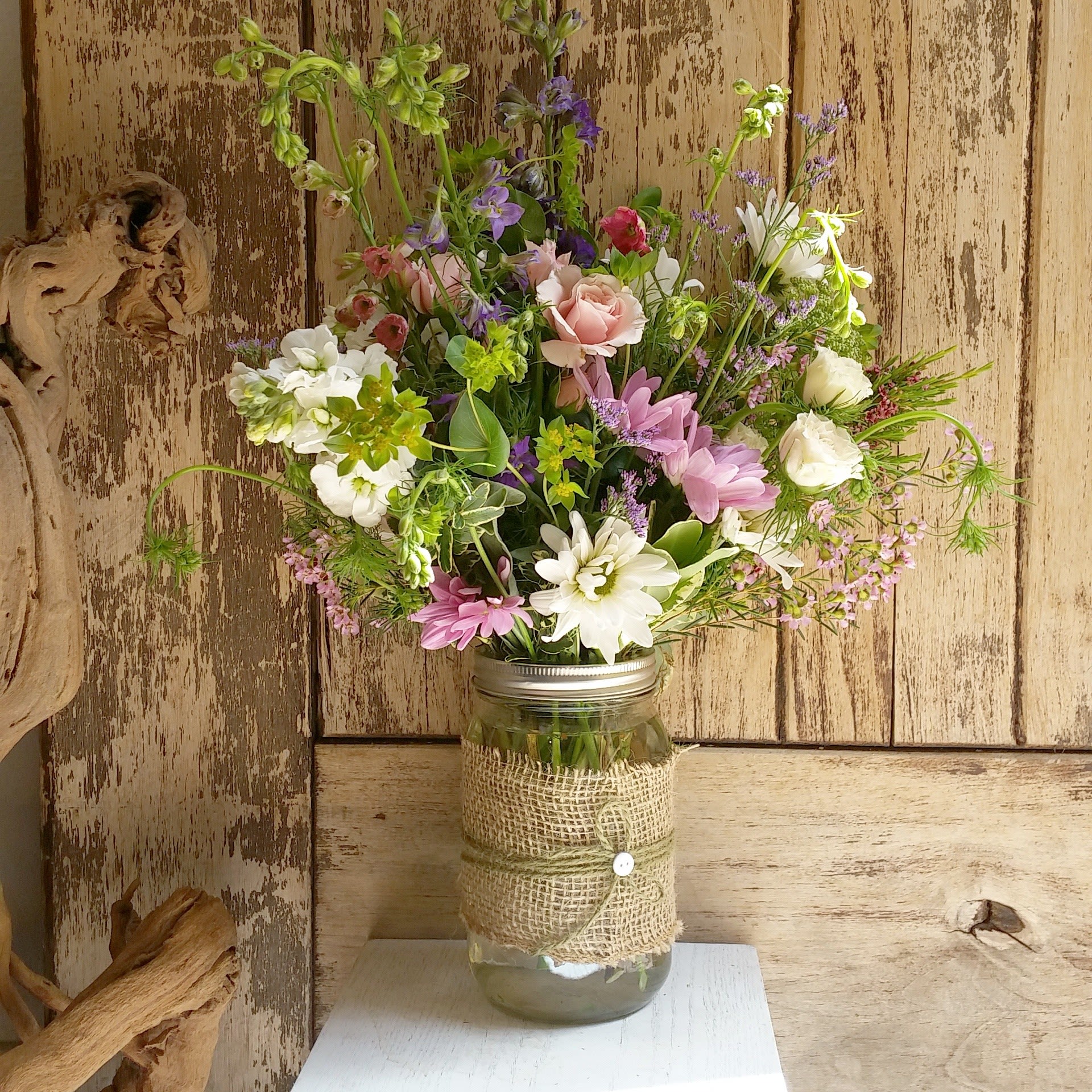 Spring Is Here - Burlap covered mason jar with daisies, larkspur and spray roses with spring accents for a fresh happy design with a cute twine bow.  PLEASE NOTE: Florist reserves the right to make modifications to the design based on seasonality and accessibility of blooms and vases. Any modifications made will honor the palette, intent, and feeling of the pictured design. Contact floral designer directly with any specifications or concerns.