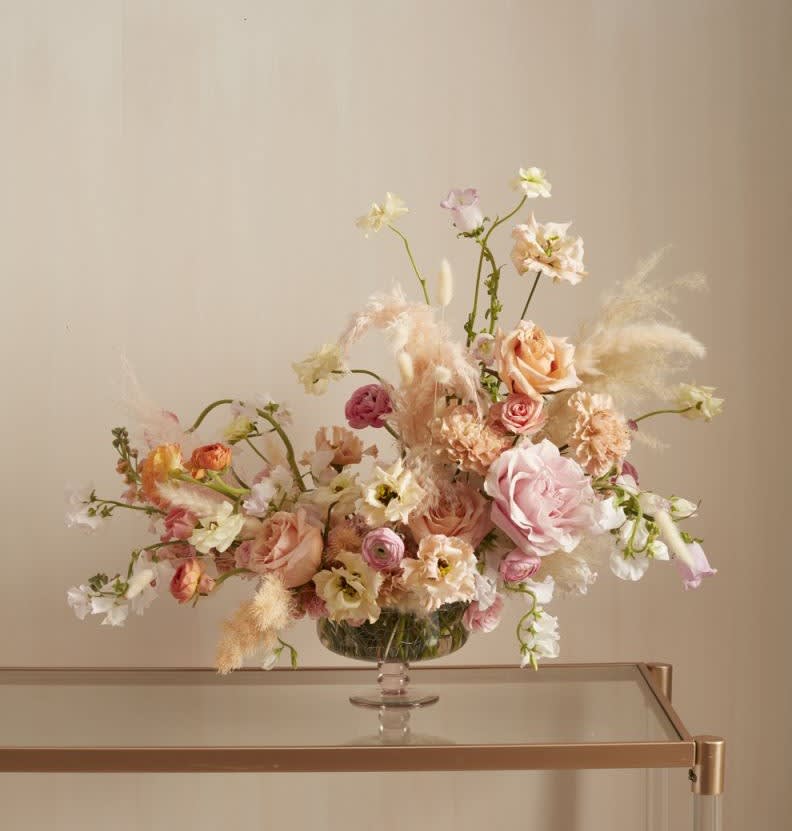 Vesna - Soft and feminine, various garden florals are arranged whimsically with fluffy pampas grass in a gorgeous glass compote bowl.