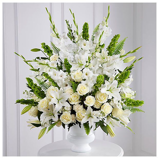 Garden of Remembrance  - This is a stunning and elegant all-white floral arrangement designed to convey peace, purity, and heartfelt remembrance. Presented in a classic white pedestal vase, it features a lush assortment of blooms beautifully complemented by rich greenery.  The arrangement includes graceful white lilies, symbolizing renewal and tranquility, alongside creamy white roses that express love, respect, and remembrance. Towering white gladiolus add height and elegance, while soft white carnations and stock flowers provide texture and fullness.  Bright green bells of Ireland and lush ferns add a vibrant contrast, enhancing the arrangement’s overall lushness. This sophisticated display is perfect for expressing sympathy, honoring a loved one, or creating an elegant centerpiece for a special occasion.