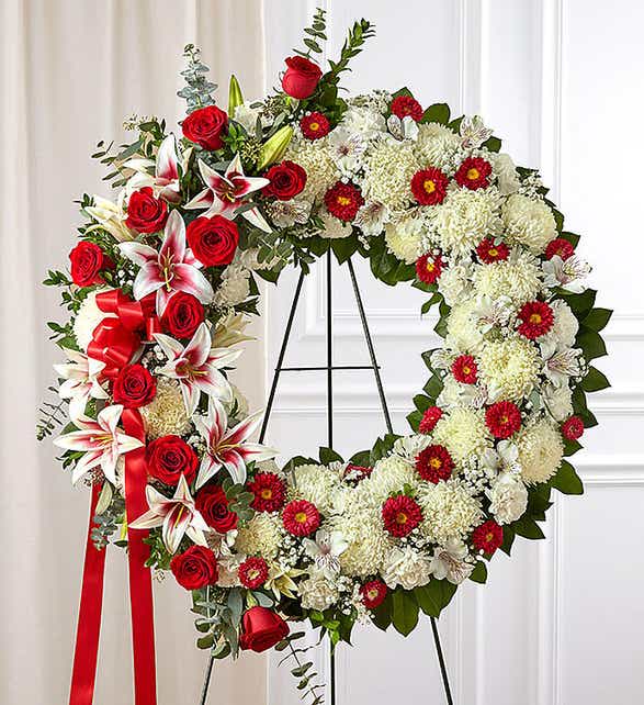 Peaceful Memory - This is a breathtaking floral wreath designed to convey love, respect, and remembrance. The circular shape symbolizes eternal life and enduring love, making it a meaningful tribute for memorial services or special occasions.  The wreath features a lush base of creamy white chrysanthemums, symbolizing purity and peace. Bright red roses, representing love and admiration, are beautifully clustered along one side, accompanied by striking red and white stargazer lilies that add elegance and fragrance. Deep red mini gerbera daisies are interspersed throughout, adding vibrant pops of color and texture.  A bold red satin ribbon cascades gracefully, enhancing the wreath’s rich, romantic aesthetic. The arrangement is completed with lush greenery, including eucalyptus, which adds depth and a natural touch. Displayed on a standing easel, this floral wreath is a heartfelt way to honor and celebrate a loved one’s life.