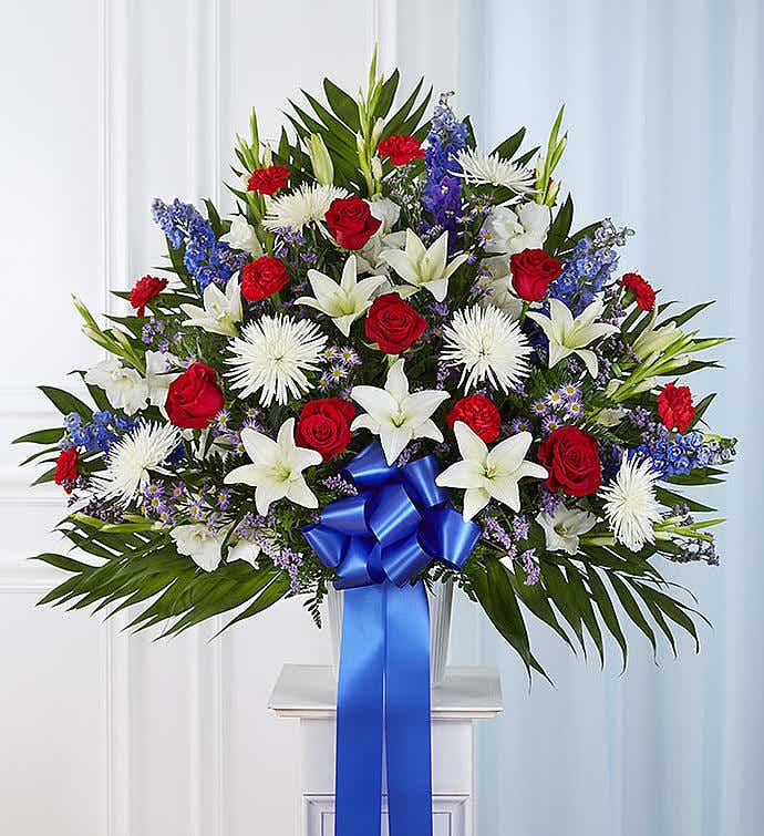 Freedom - This is a striking and patriotic floral arrangement, beautifully designed with a classic red, white, and blue color palette. It conveys a sense of honor, respect, and remembrance, making it a perfect tribute for memorial services, patriotic events, or to honor someone special.  The arrangement features vibrant red roses and carnations, symbolizing love and admiration. Elegant white lilies and spider mums represent purity and peace, while rich blue delphiniums and purple filler flowers add depth and a touch of serenity.  Lush palm leaves frame the arrangement, providing structure and enhancing the vibrant blooms. A bold royal blue satin bow cascades down the center, tying the theme together with an elegant finish. Presented in a traditional white urn, this arrangement makes a meaningful and dignified statement for any occasion. PICTURE IS STANDAR SIZE