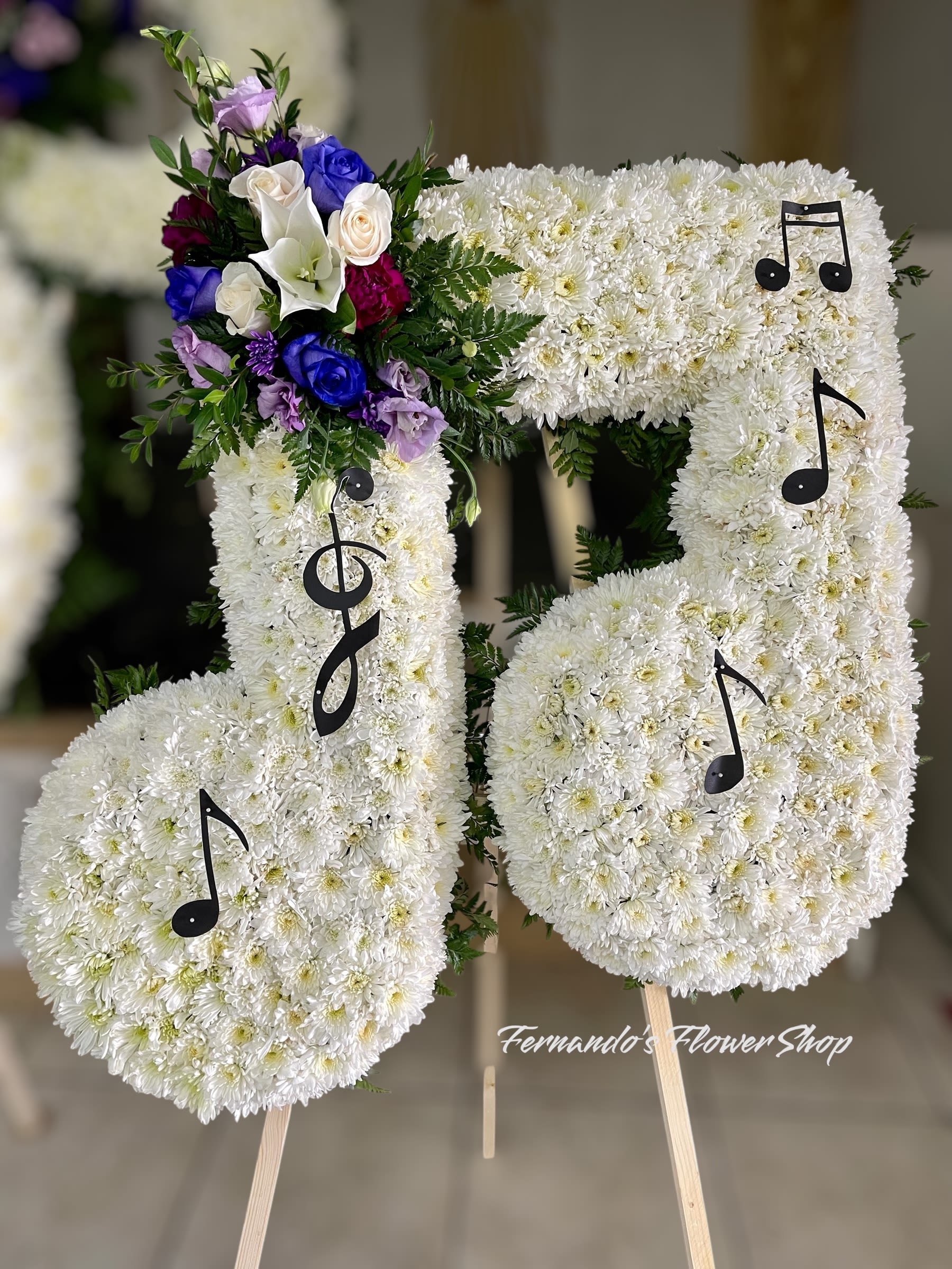 Music Note Music Note - This is a stunning musical-themed floral arrangement designed in the shape of two musical notes. The notes are crafted with tightly packed white chrysanthemums, symbolizing purity, peace, and remembrance. Black musical symbols, including treble and eighth notes, are placed on the arrangement, enhancing the musical theme. A vibrant floral accent adorns the top left side, featuring purple, blue, red, and cream roses, along with white calla lilies and lush greenery. This pop of color adds contrast and elegance to the arrangement. This type of tribute is ideal for honoring someone with a deep love for music, representing their passion and the lasting melody they leave behind.
