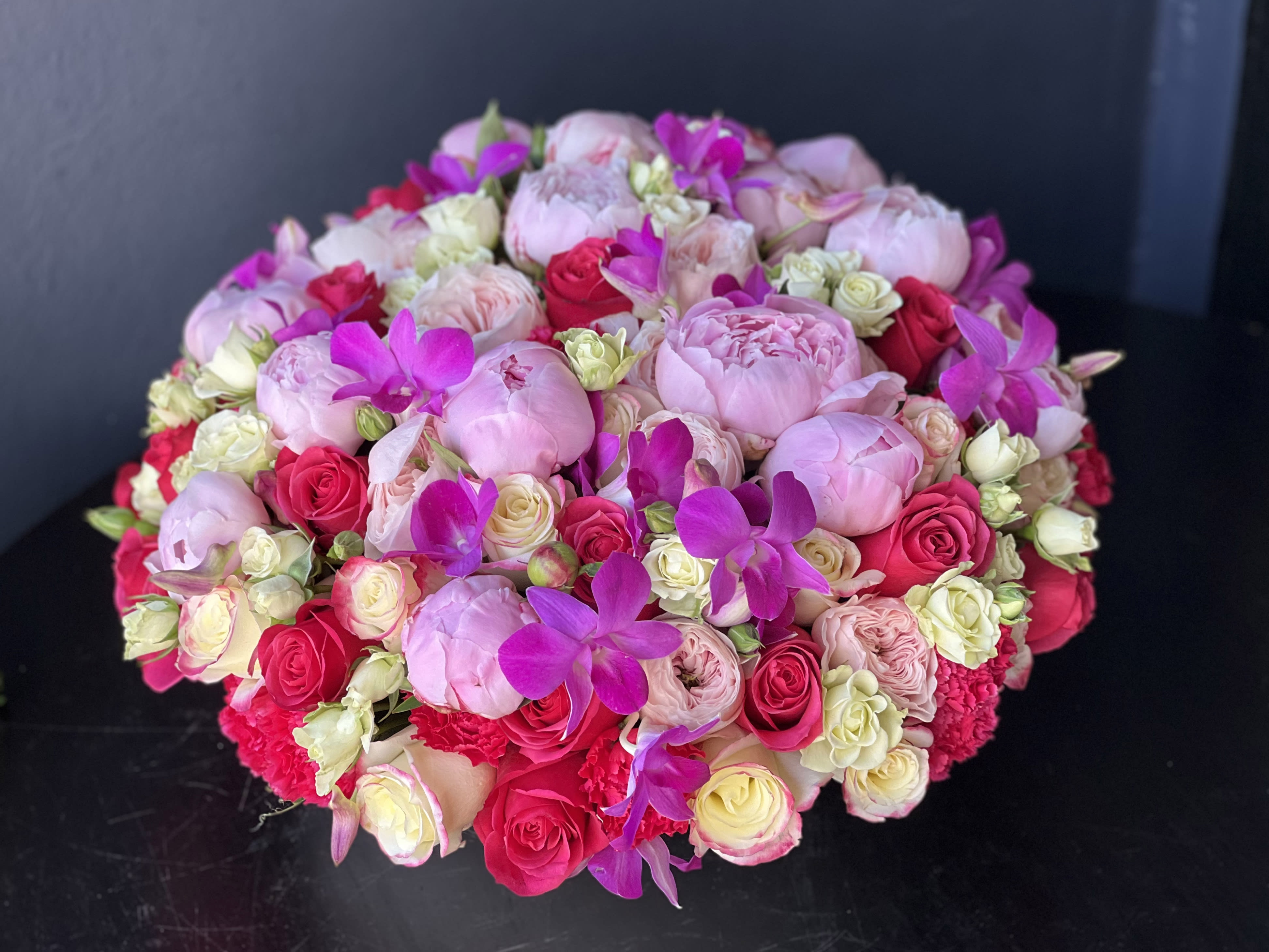 BLOOMING LOVE  - Details:  Sophisticated flower design in cement bowl with hot pink and pink roses, spray roses, carnations, dendrobium orchids, peonies, greenery and mixed seasonal flowers.