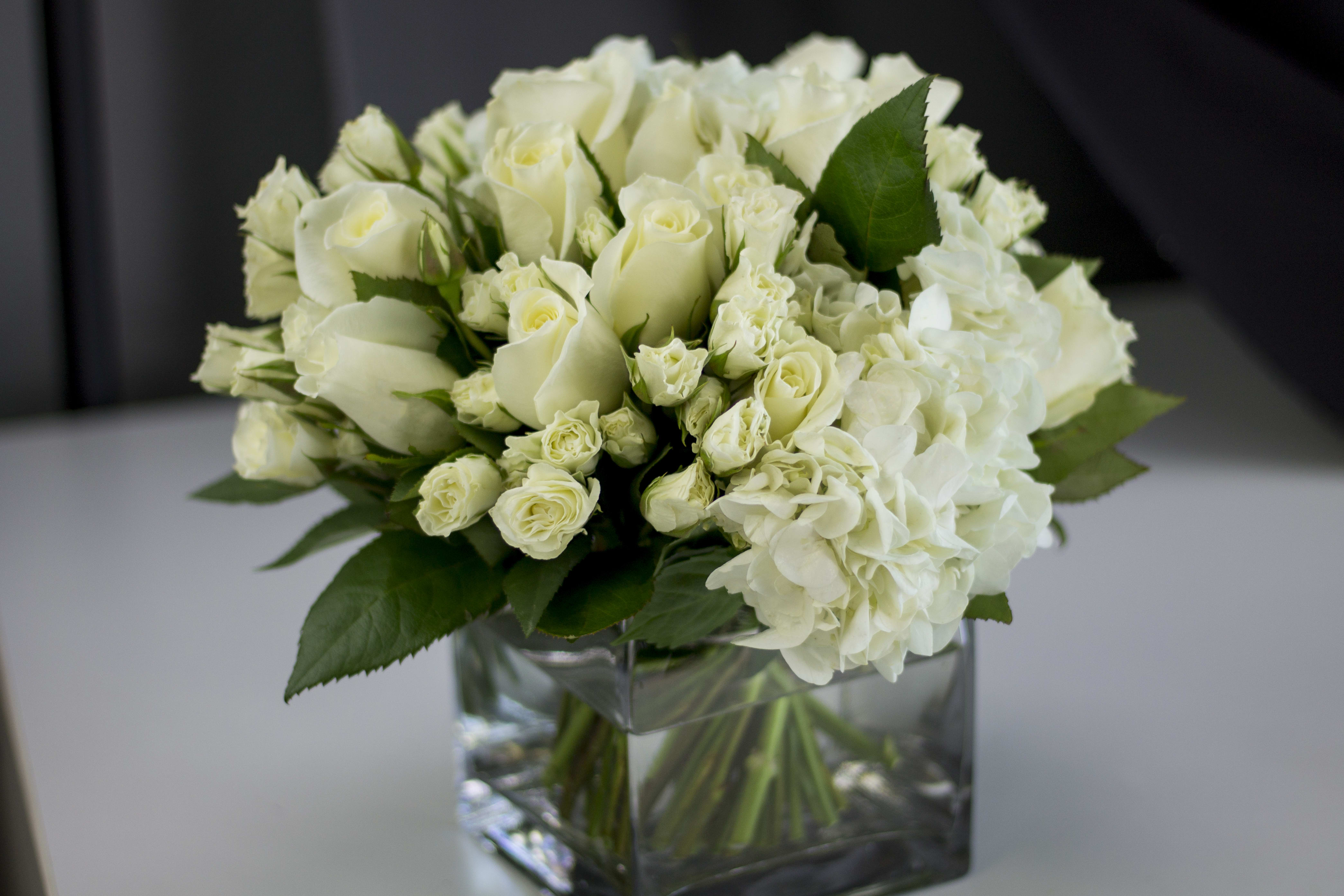 All in white arrangement - Classic mix of white roses, spray roses and hydrangeas. Arranged into square clear vase