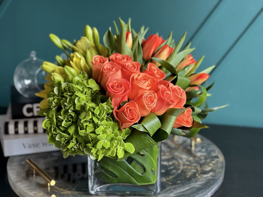 Sunset  - Bright sunny design with full of flowers, true orange roses, tulips, yellow asiatic lilies and green hydrangeas based in cube vase.   