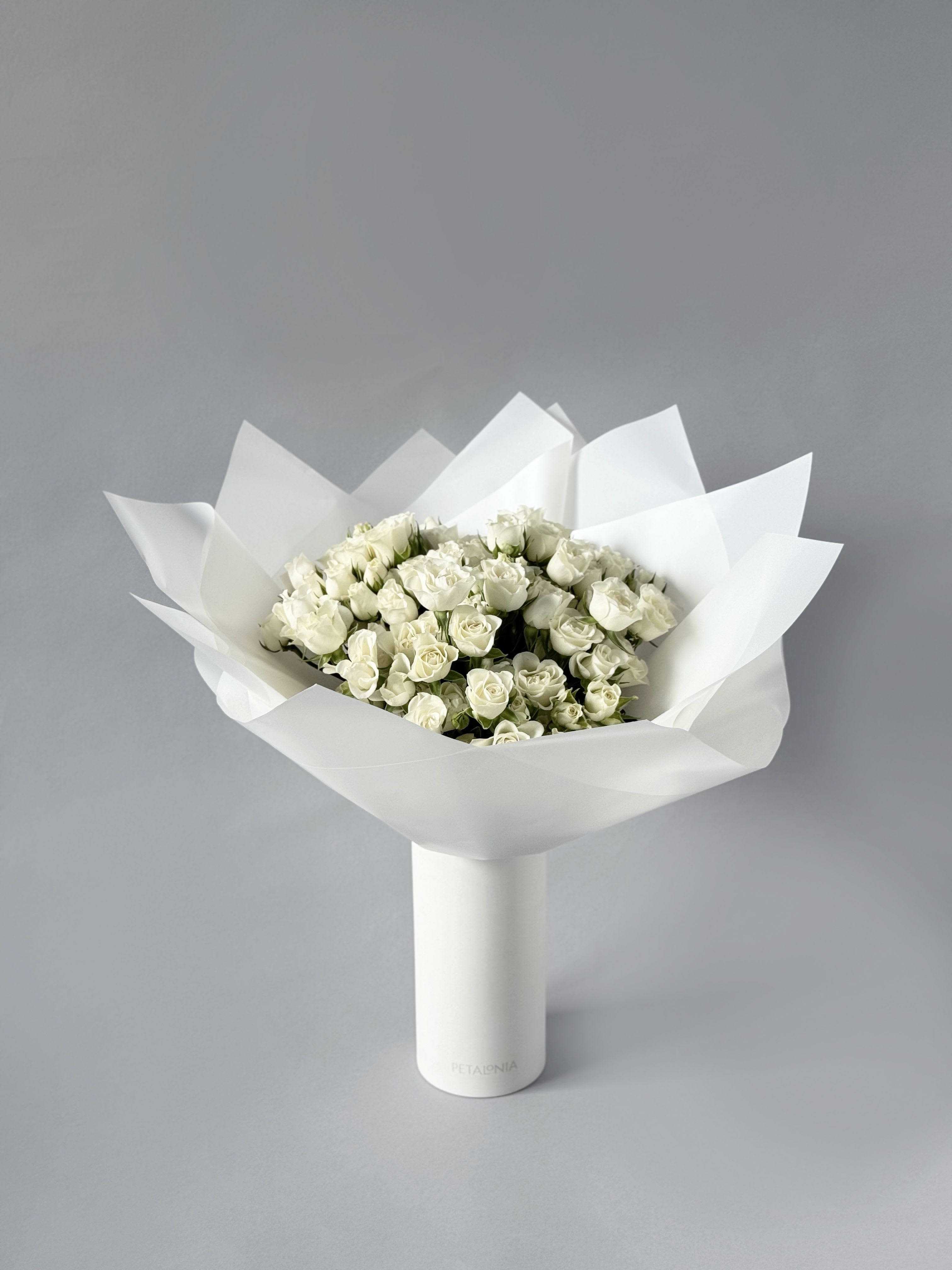 White Mini Roses in a Signature Petalonia Vase and Tote - Experience pure elegance with Petalonia's mini roses—beautifully arranged in our signature vase, wrapped in trendy Korean paper, and delivered in a stylish Petalonia tote, adding the perfect touch of luxury to any occasion.  Vase Dimensions 3.75” x 8.25” 