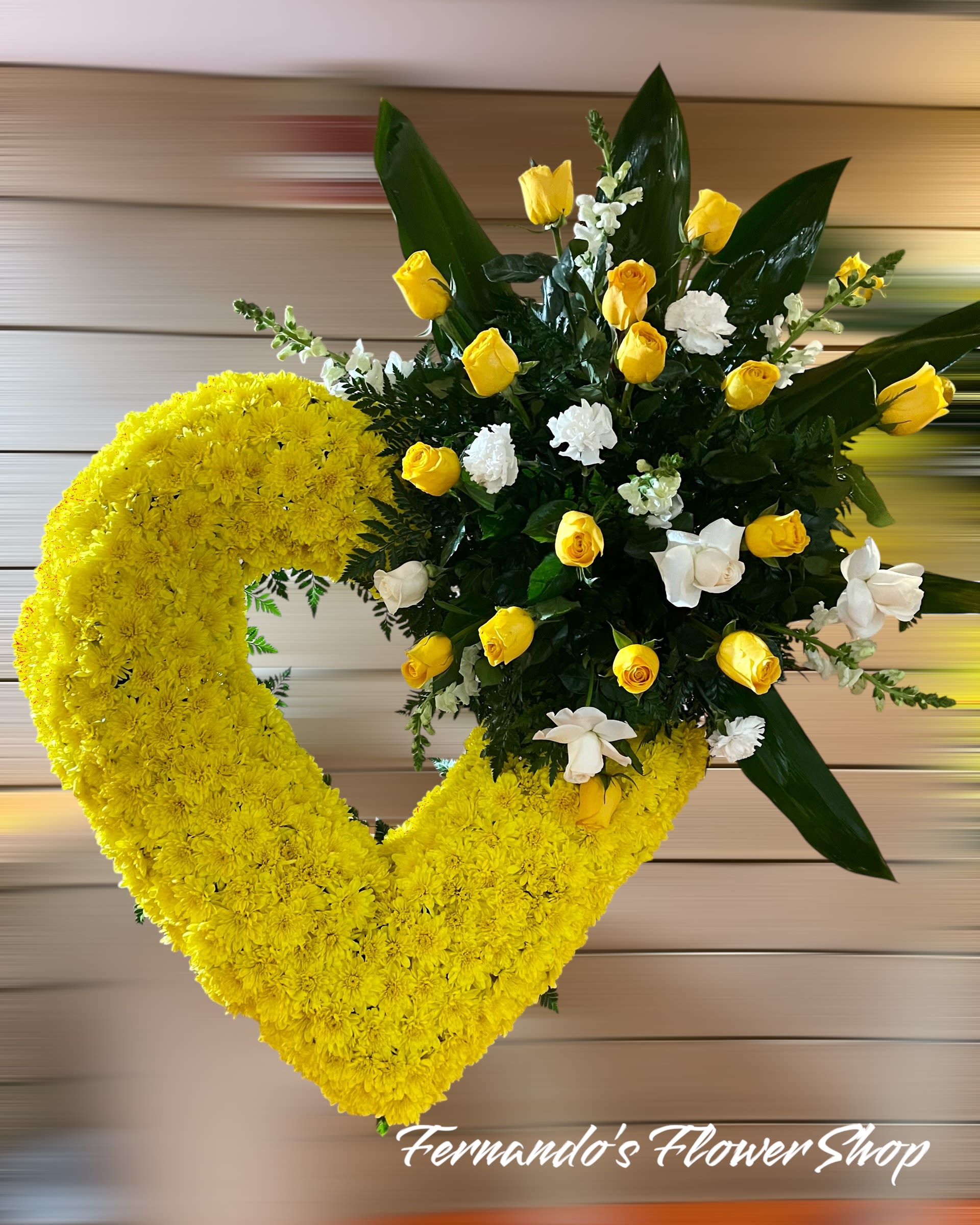 Beaty in Yellow - This is a vibrant heart-shaped floral arrangement featuring bright yellow and white flowers. The heart is densely covered with yellow chrysanthemums, symbolizing friendship, joy, and remembrance. A striking floral spray emerges from the upper right, consisting of yellow roses, white roses, and white carnations, complemented by lush green foliage and large tropical leaves. This arrangement conveys a message of love, respect, and warmth, making it a beautiful tribute for a memorial or funeral service. The bottom of the image features the text “Fernando’s Flower Shop,” indicating the florist.