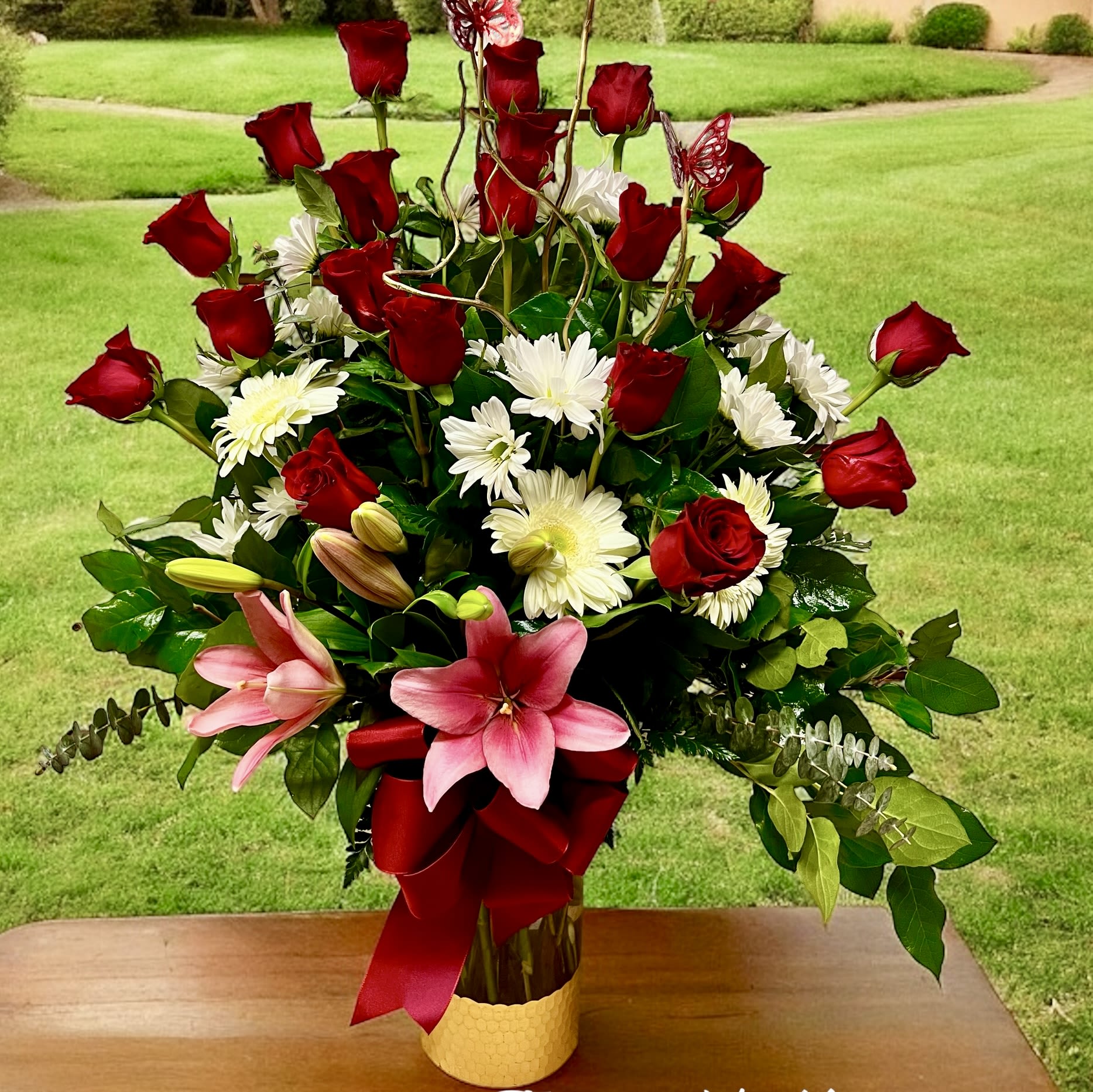 Mi Amore - This is a stunning and elegant floral arrangement in a golden vase, featuring a vibrant mix of red roses, white daisies, and pink lilies. The red roses, symbolizing love and passion, are prominently arranged at the top, while the white daisies add purity and brightness. The pink lilies bring a touch of grace and femininity to the arrangement. Lush green foliage, including eucalyptus leaves, provides contrast and fullness. A large red ribbon bow at the base enhances the overall presentation, making this bouquet perfect for a romantic gesture, celebration, or tribute. The outdoor background adds a fresh, natural touch to the display.