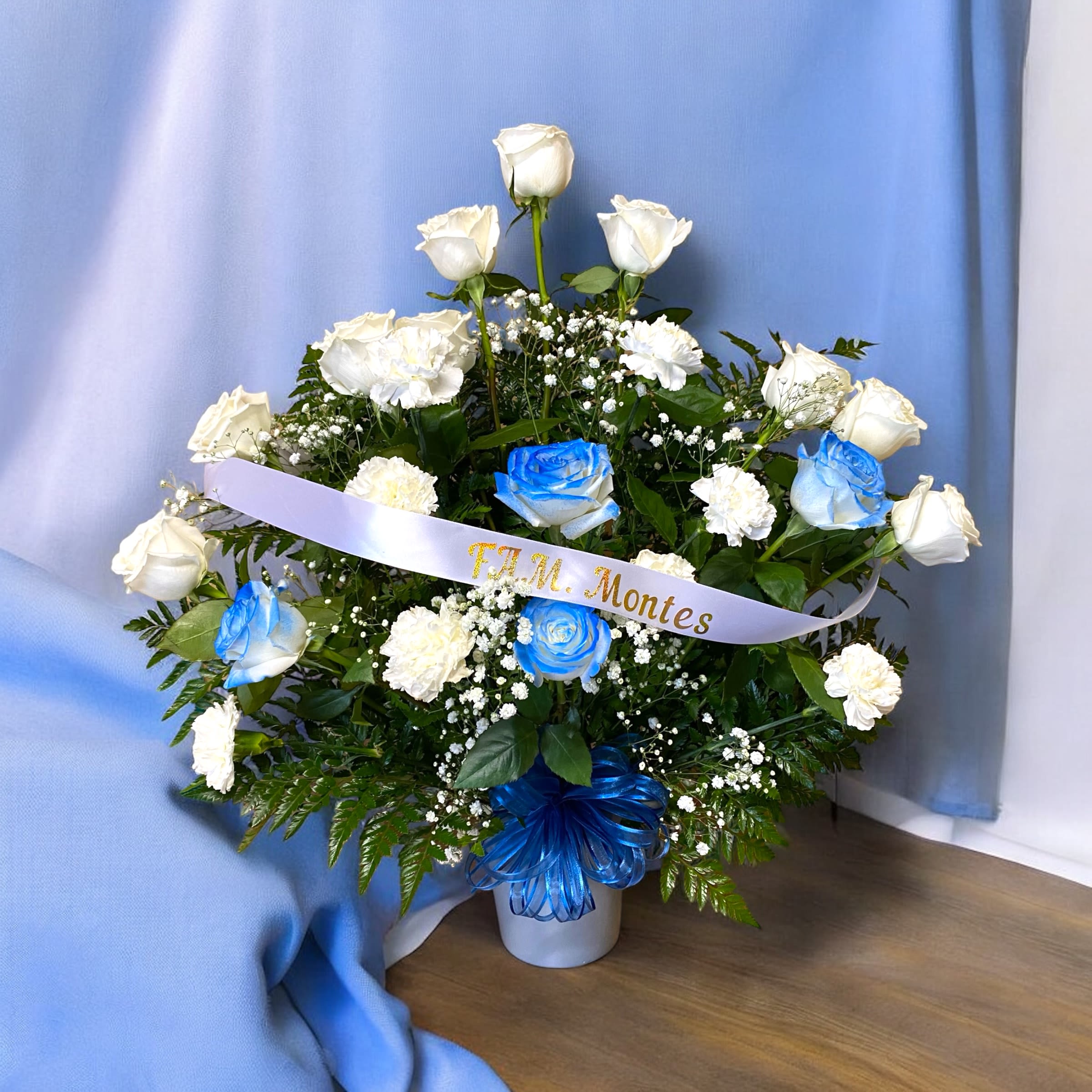 Elegant Blue and White Funeral Floral Arrangement - Honor the memory of a loved one with this elegant blue and white funeral floral arrangement. This thoughtfully designed tribute features a harmonious blend of white roses, carnations, and baby’s breath, symbolizing purity and remembrance. Beautifully accented with striking blue roses, the arrangement conveys a sense of peace and serenity.    A delicate white ribbon, personalized with golden lettering, adds a heartfelt touch, making this arrangement a fitting expression of sympathy and support. Adorned with lush greenery and presented in a white vase, it serves as a dignified tribute for funeral services or memorial ceremonies.    This floral arrangement is a respectful and graceful way to convey your condolences and honor the memory of someone dear.