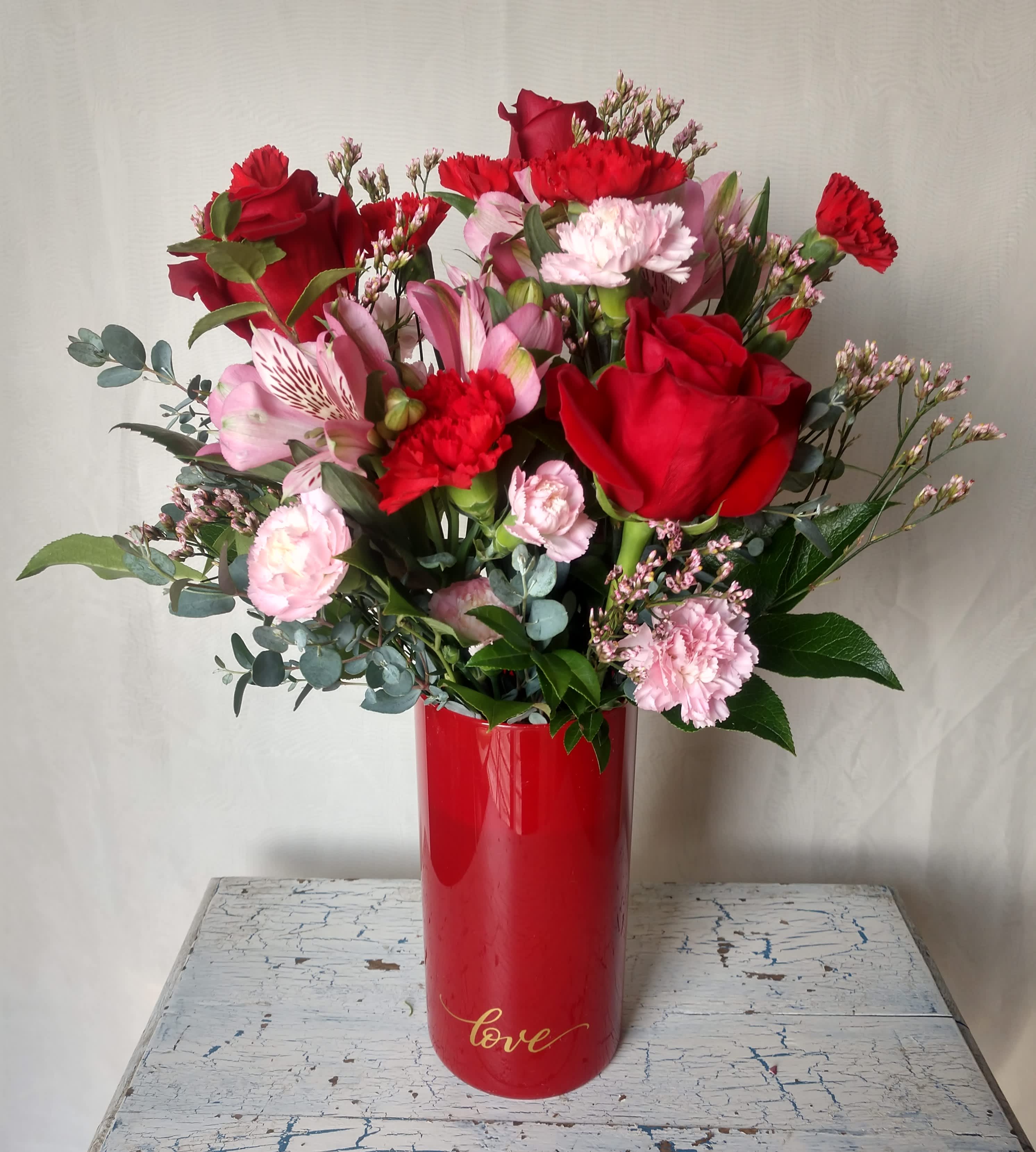Blooming Love - Sweet and romantic mix of alstroemeria, roses and carnations with assorted greens and filler arranged in beautiful red glass cylinder vase with the word love painted in gold.